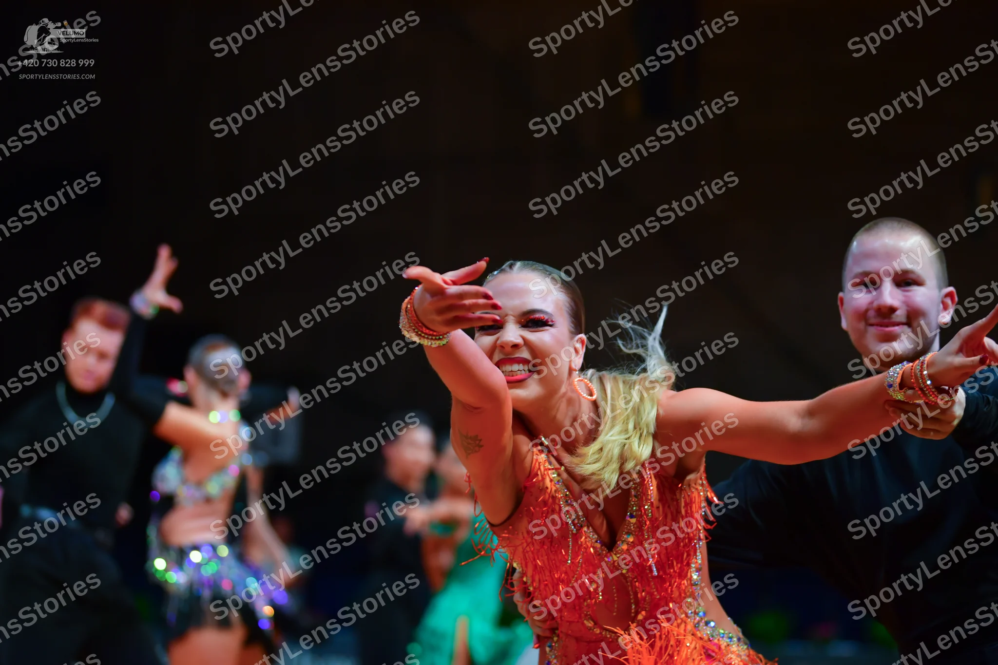 Dancesport competitor: Foreground female dancer: orange-red Latin dance dress with sequins and fringe