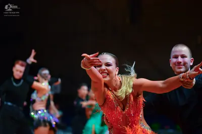 Dancesport competitor: Foreground female dancer: orange-red Latin dance dress with sequins and fringe