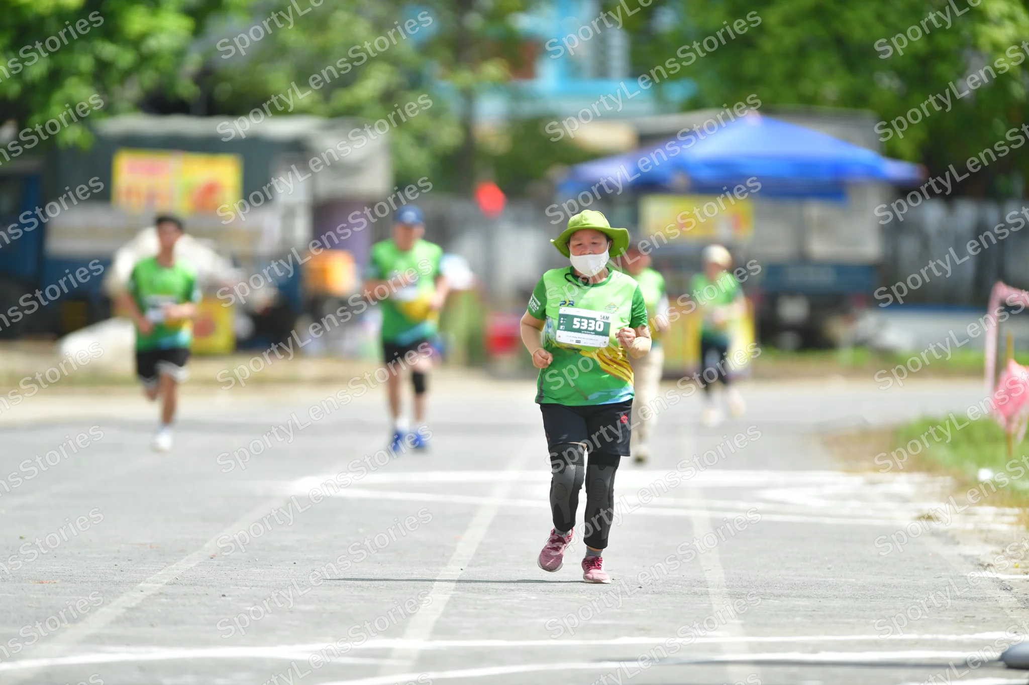 Marathon runner #5320: Runner 1: green jersey, black leggings, pink shoes, green sun hat, white face ma