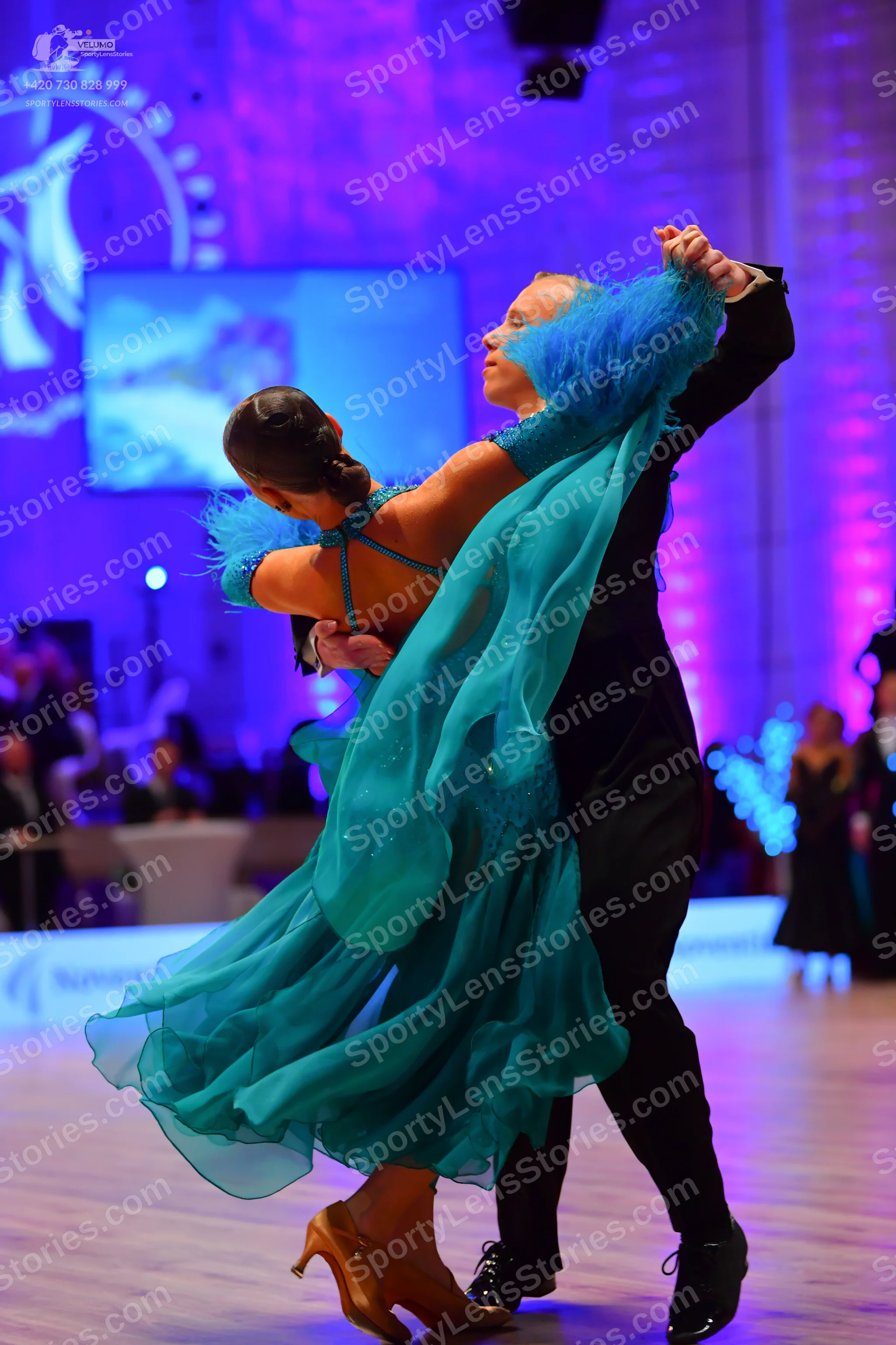 Dancesport competitor: Female dancer: teal flowing dress with a multi-layered skirt and feathery/textur