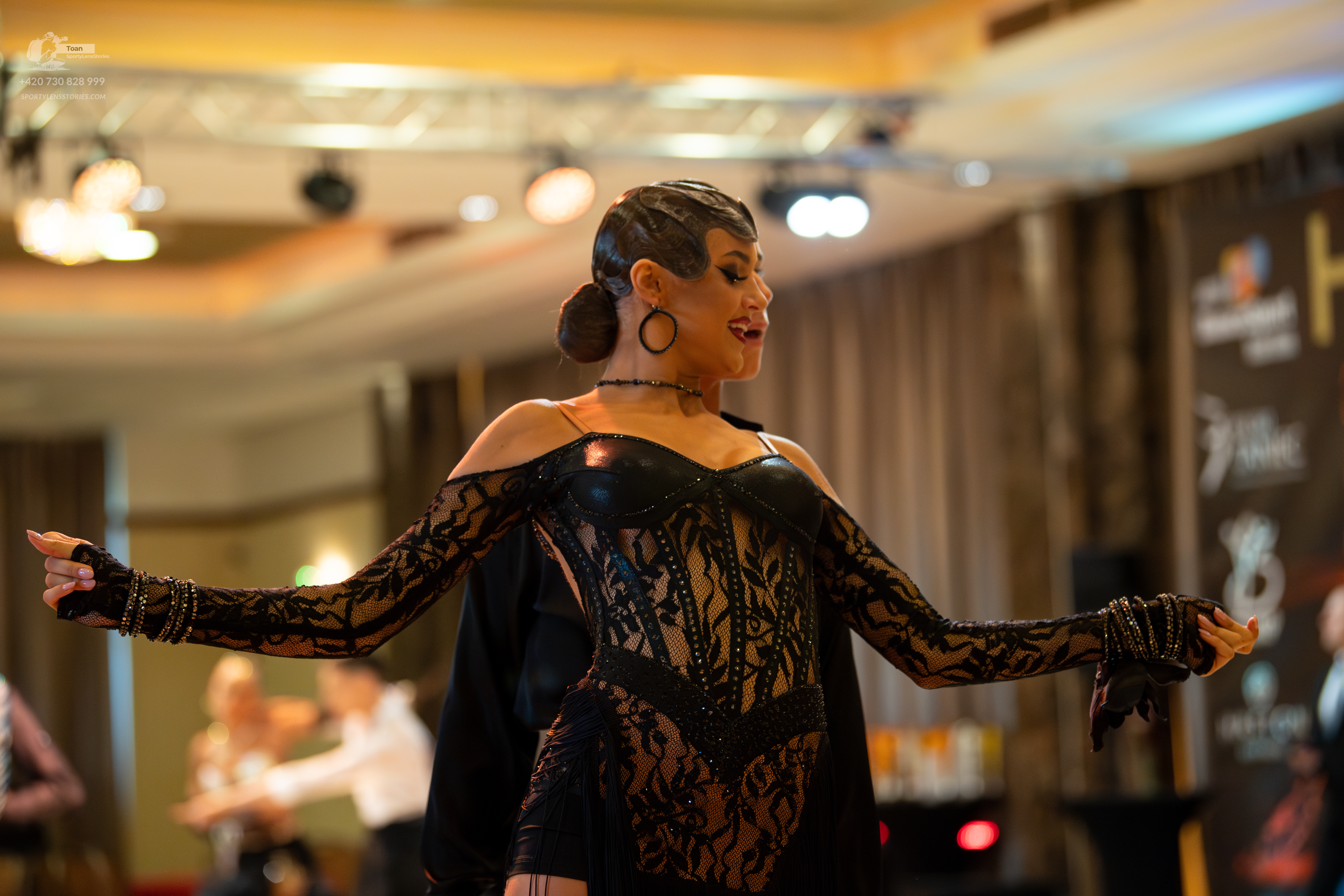 Dancesport competitor: Female dancer (foreground): black Latin dance costume featuring long floral lace