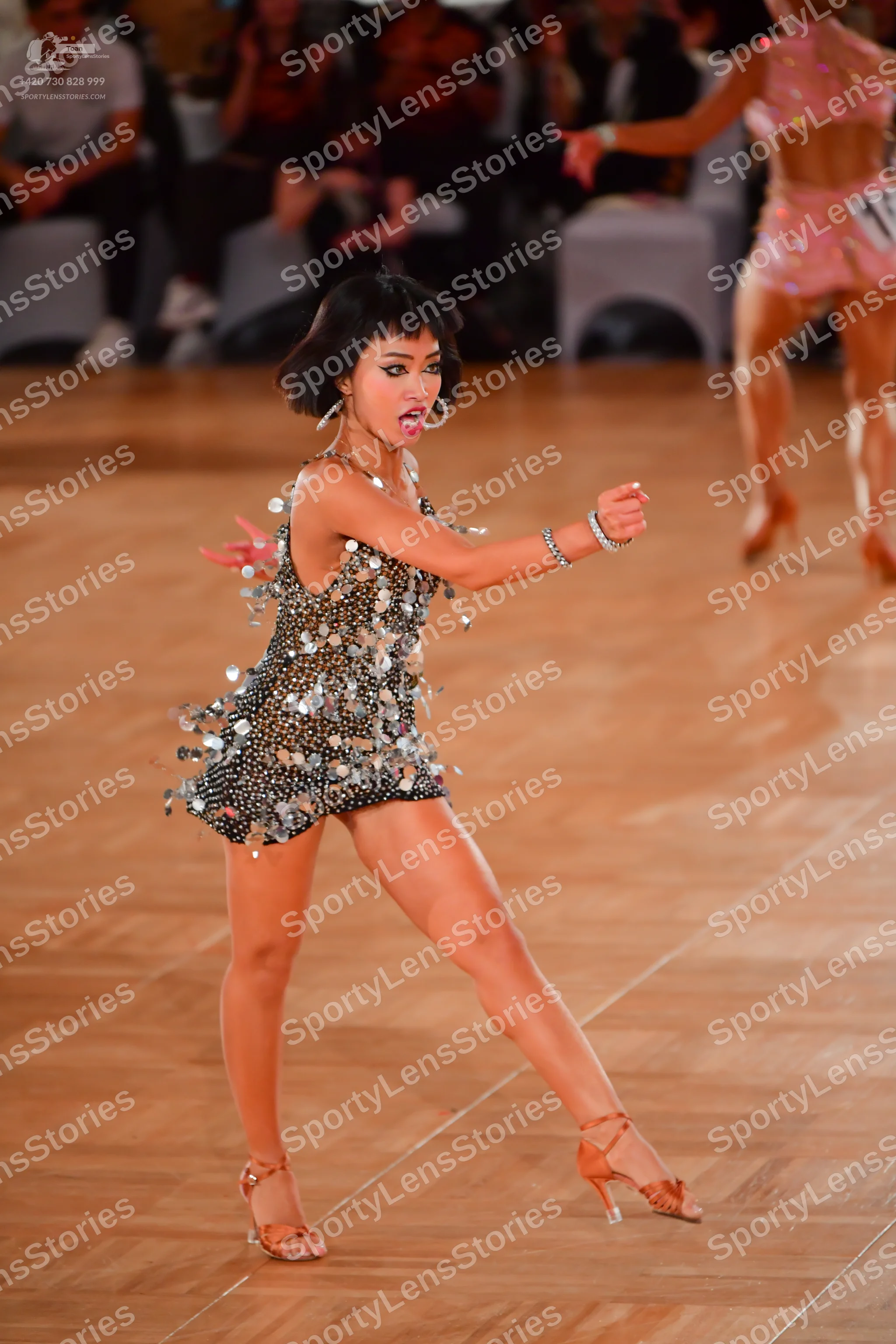 Dancesport competitor: Female dancer in foreground: dark grey/black Latin dance dress covered in large