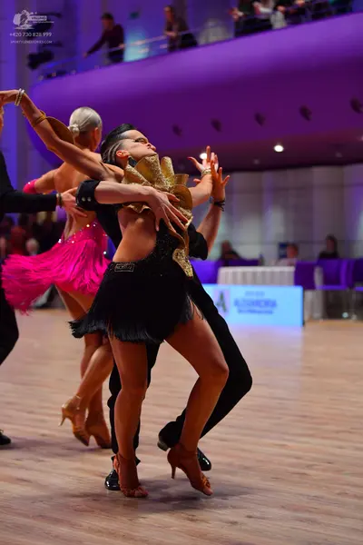 Dancesport competitor: Female dancer in foreground: black Latin dress with a large, prominent gold sequ
