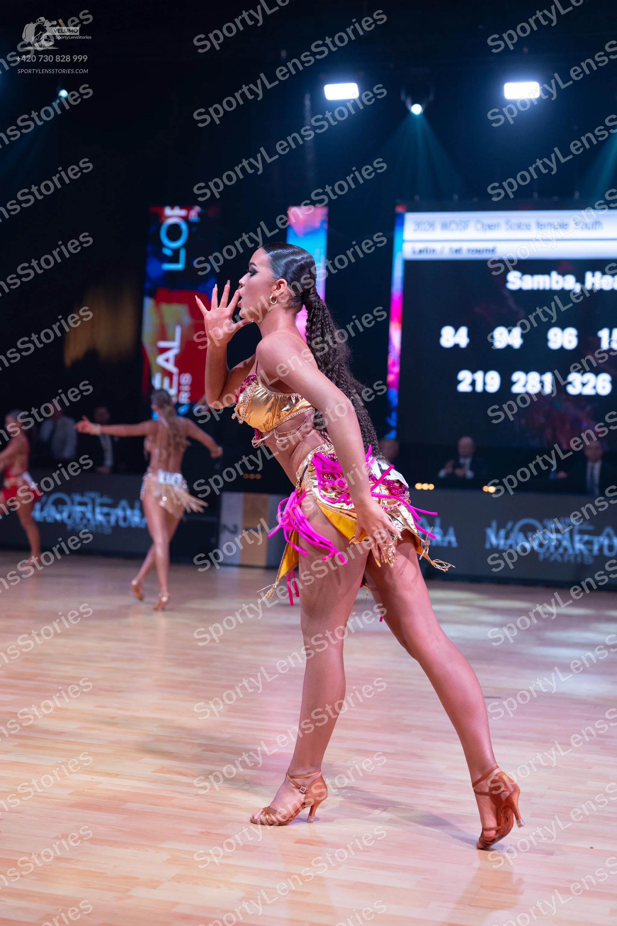 Dancesport competitor: Female dancer in foreground: gold sequined bra top and gold skirt with pink frin