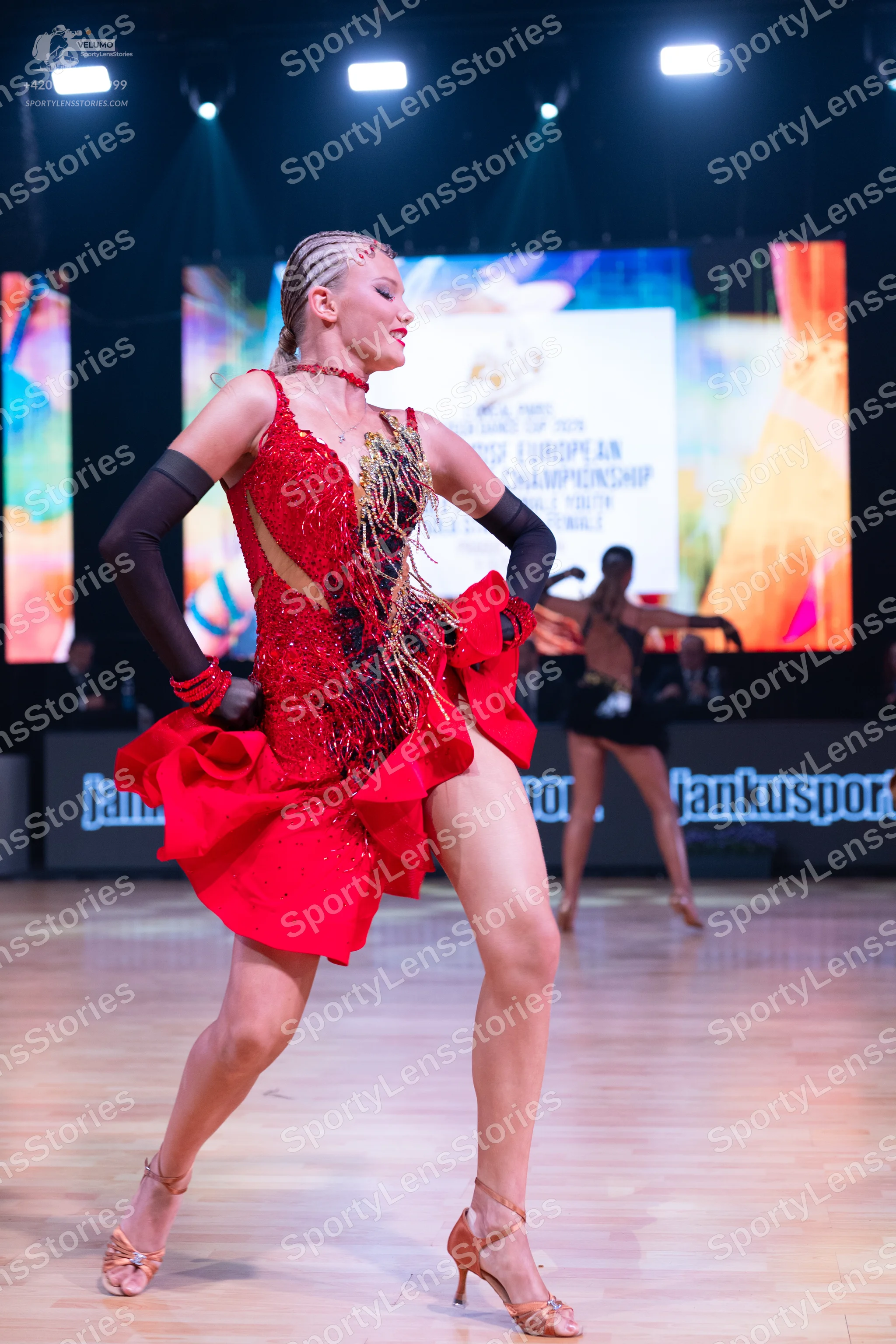 Dancesport competitor: Female dancer in foreground: red latin dress with sequins and ruffles, paired wi