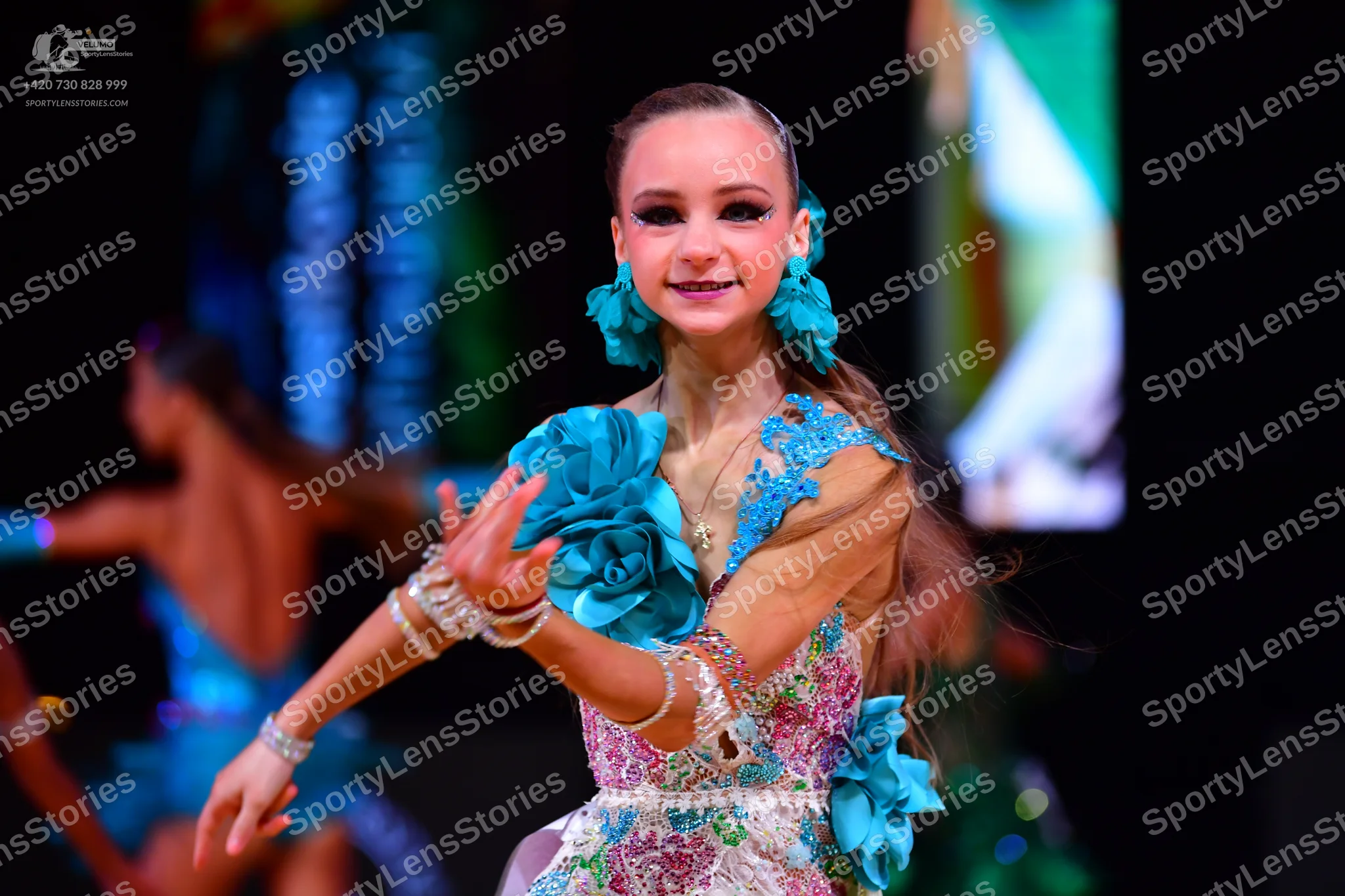 Dancesport competitor: Female dancer in foreground: teal and white Latin dance dress with large 3D teal
