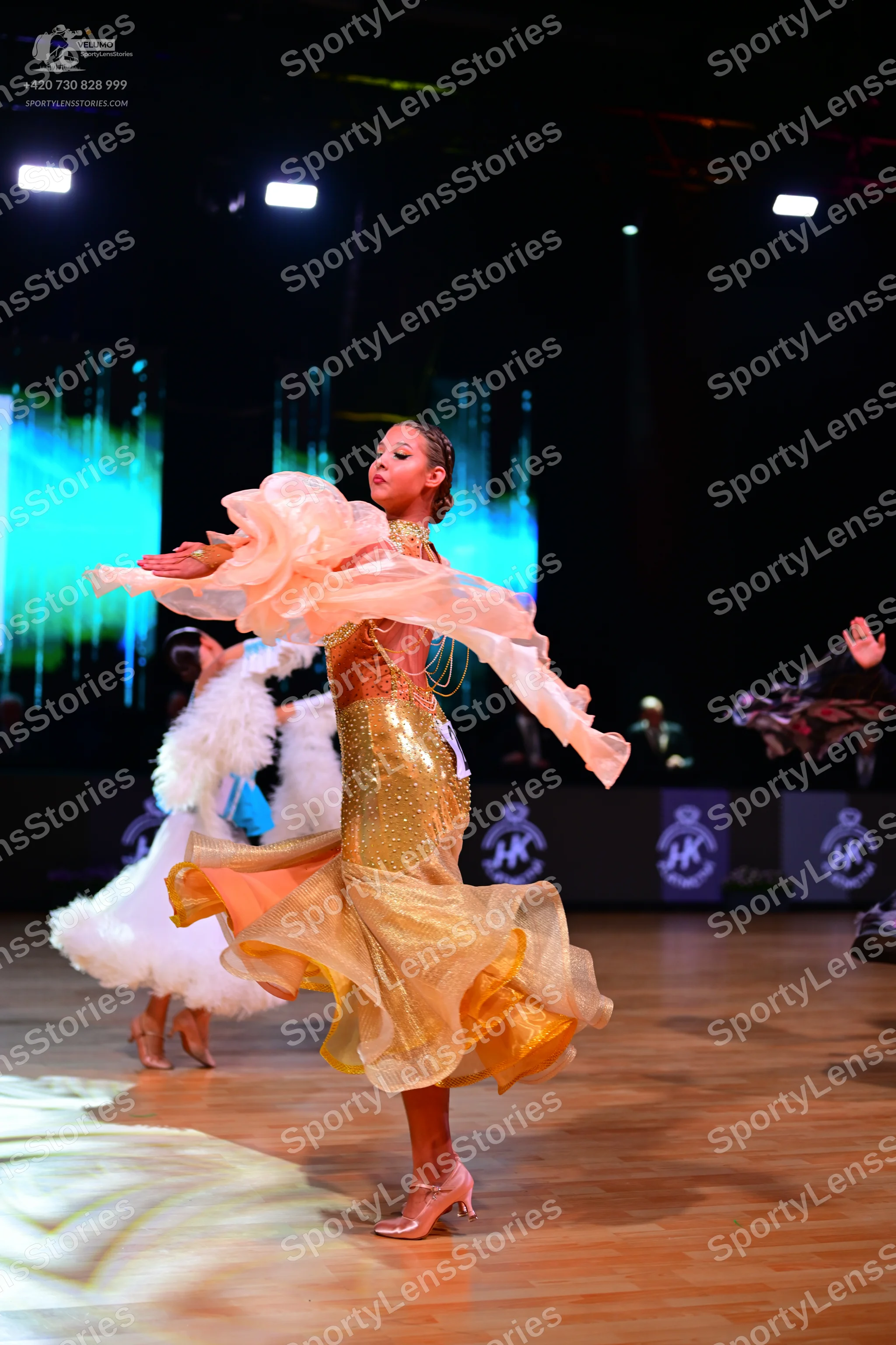 Dancesport competitor: Female dancer (foreground): Gold/amber Latin dance dress with shimmering, possib