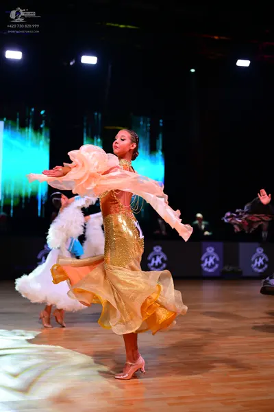 Dancesport competitor: Female dancer (foreground): Gold/amber Latin dance dress with shimmering, possib