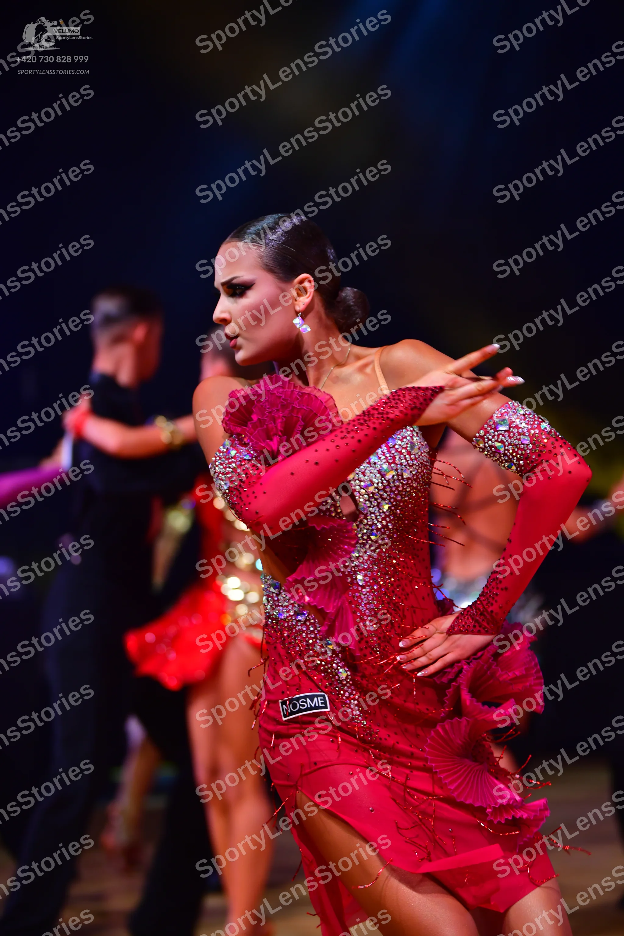Dancesport competitor: Female dancer in foreground: red latin dress with sequins, ruffles, and long sle