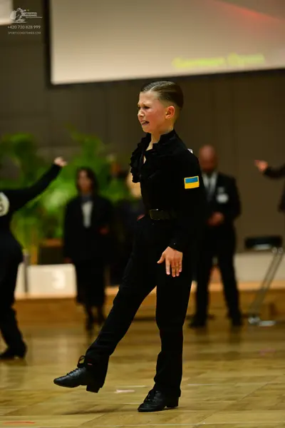 Dancesport competitor: Male dancer: black long-sleeved top with a ruffled collar and a Ukrainian flag p
