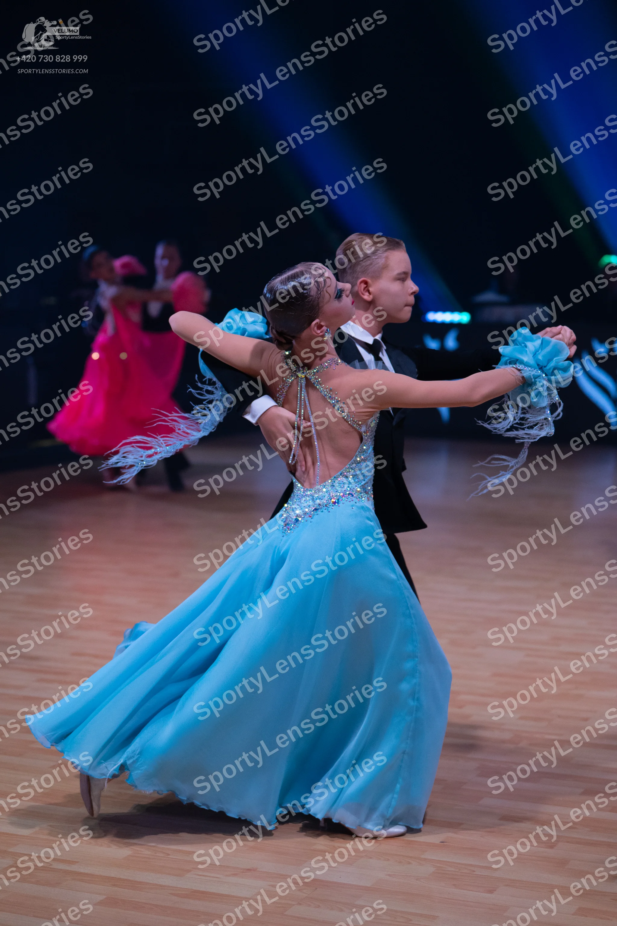 Dancesport competitor: Female dancer in foreground: light blue ballroom gown with a flowing skirt, a se
