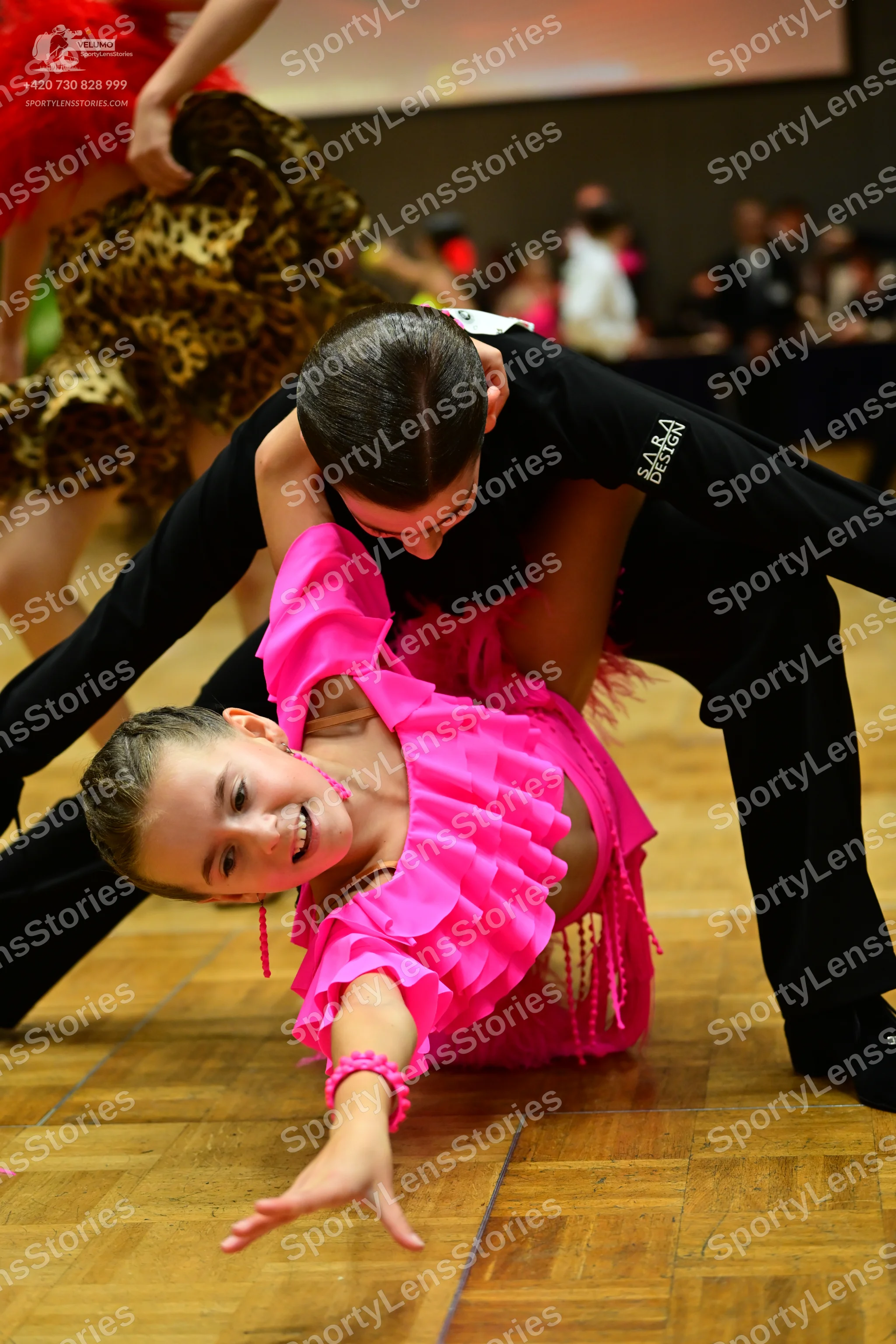 Dancesport competitor: Foreground female dancer: bright pink Latin dance dress with multiple ruffles on