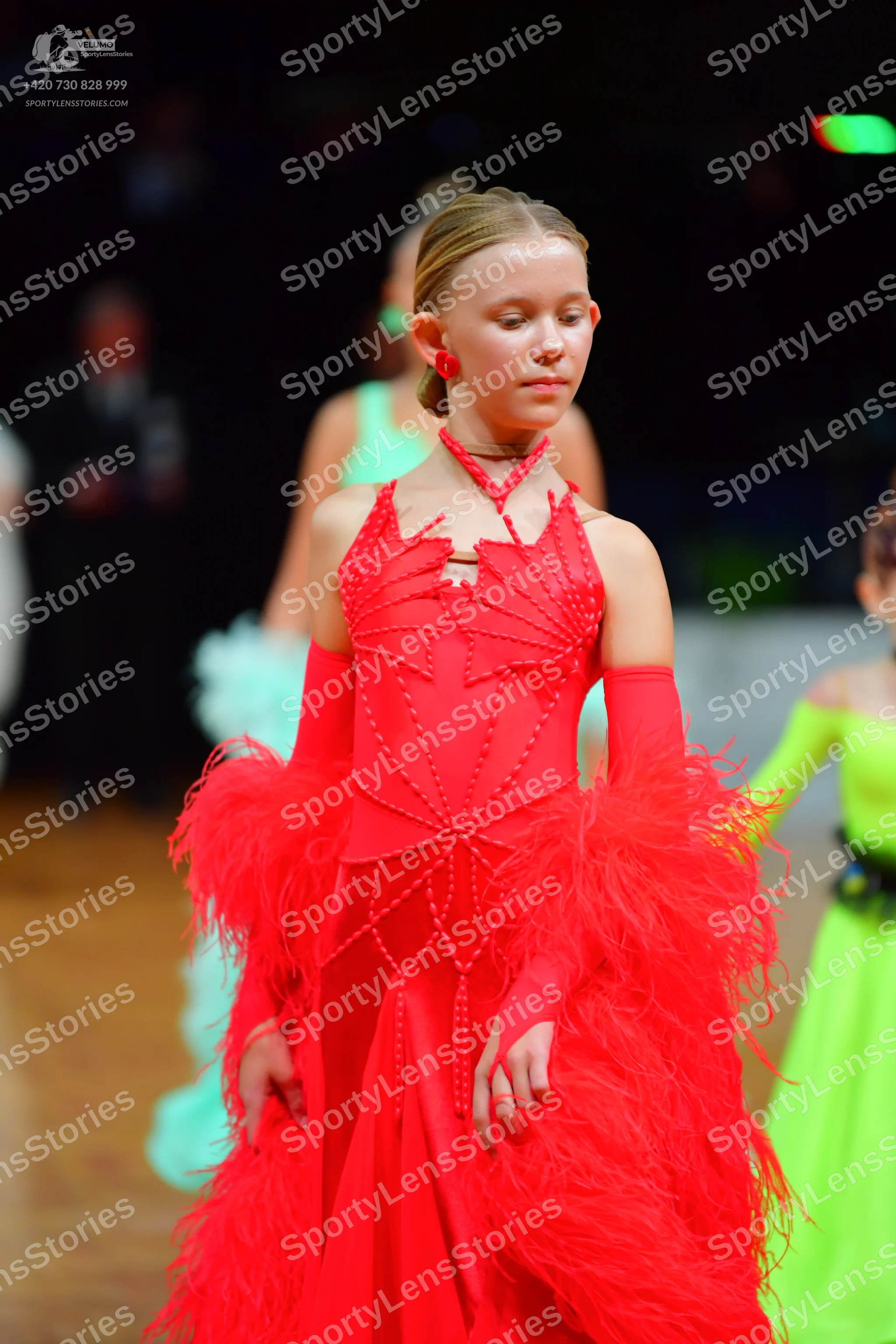 Dancesport competitor: Female dancer in foreground: red Latin dress with red beadwork and large red fea
