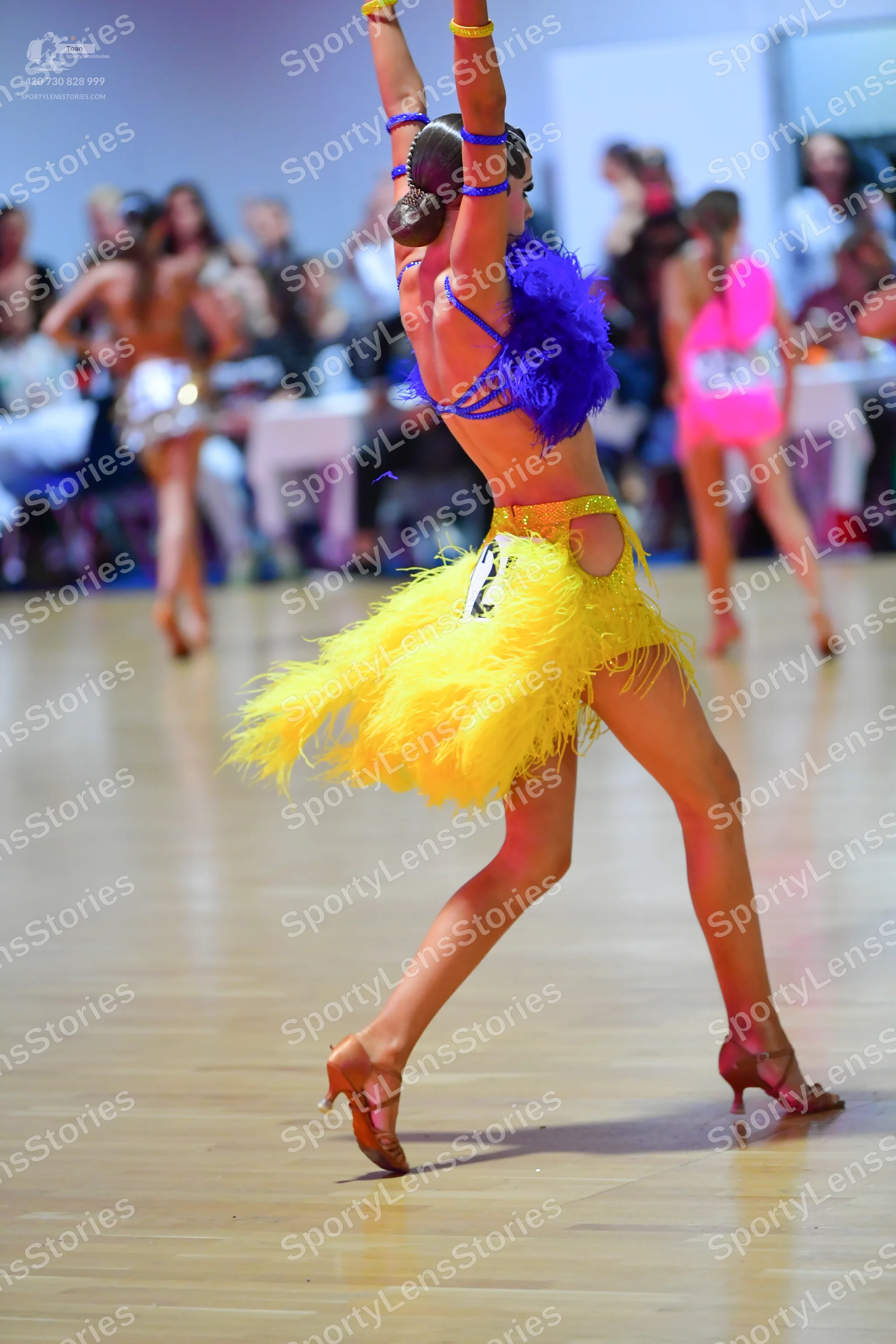 Dancesport competitor: Foreground dancer: yellow Latin dress with fringe and thin blue straps, featurin