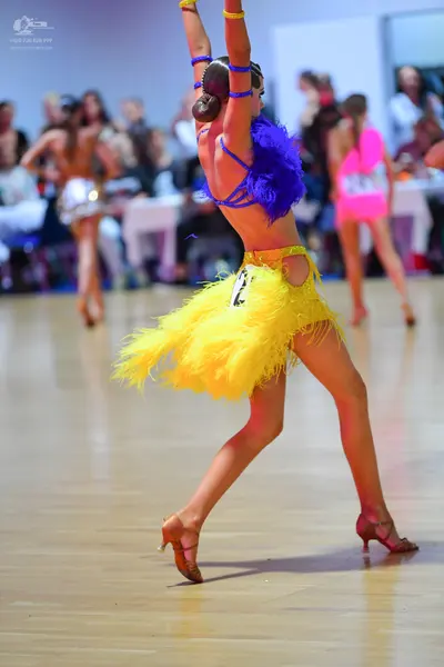 Dancesport competitor: Foreground dancer: yellow Latin dress with fringe and thin blue straps, featurin
