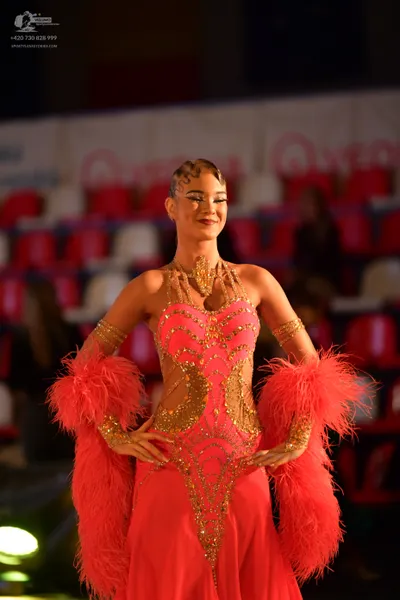 Dancesport competitor: Female dancer: red/coral dress with gold sequin and bead embellishments on a mes