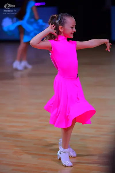Dancesport competitor: Female dancer in foreground: bright neon pink sleeveless Latin dress with a flar