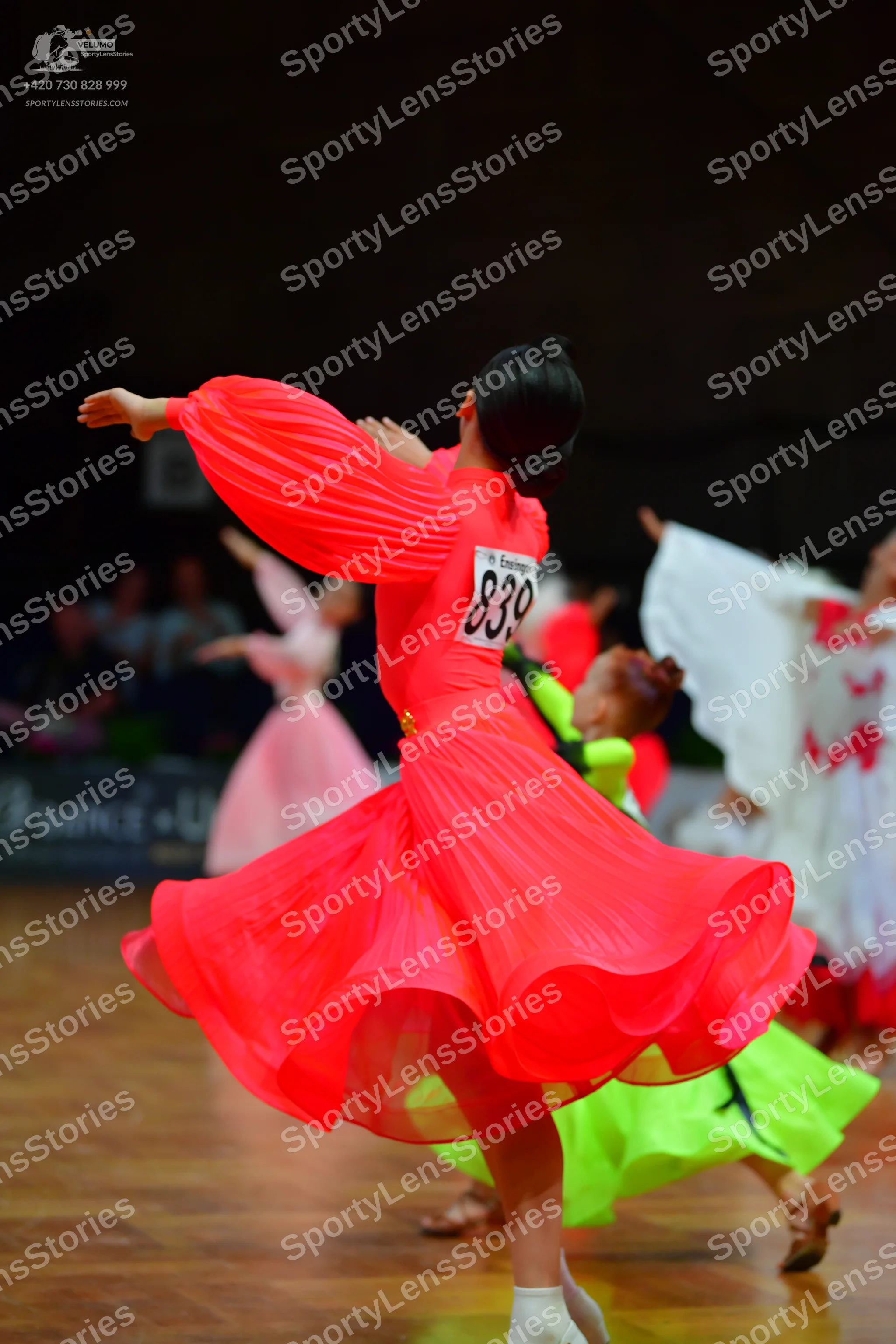 Dancesport competitor: Female dancer in foreground: reddish-pink long-sleeved ballroom dress with pleat