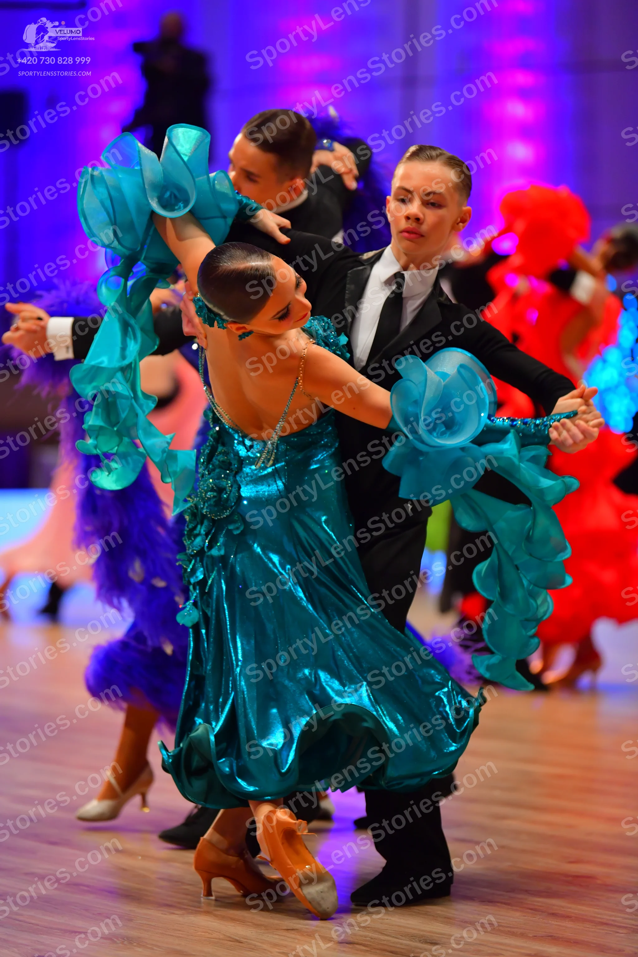Dancesport competitor: Female dancer in foreground: teal metallic ballroom dress with large, flowing, r