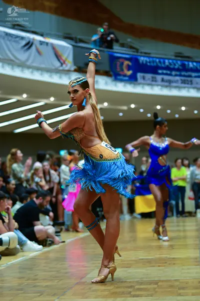 Dancesport competitor: Female dancer 1: tan Latin dress with blue fringe, gold/yellow sequin patterns, 