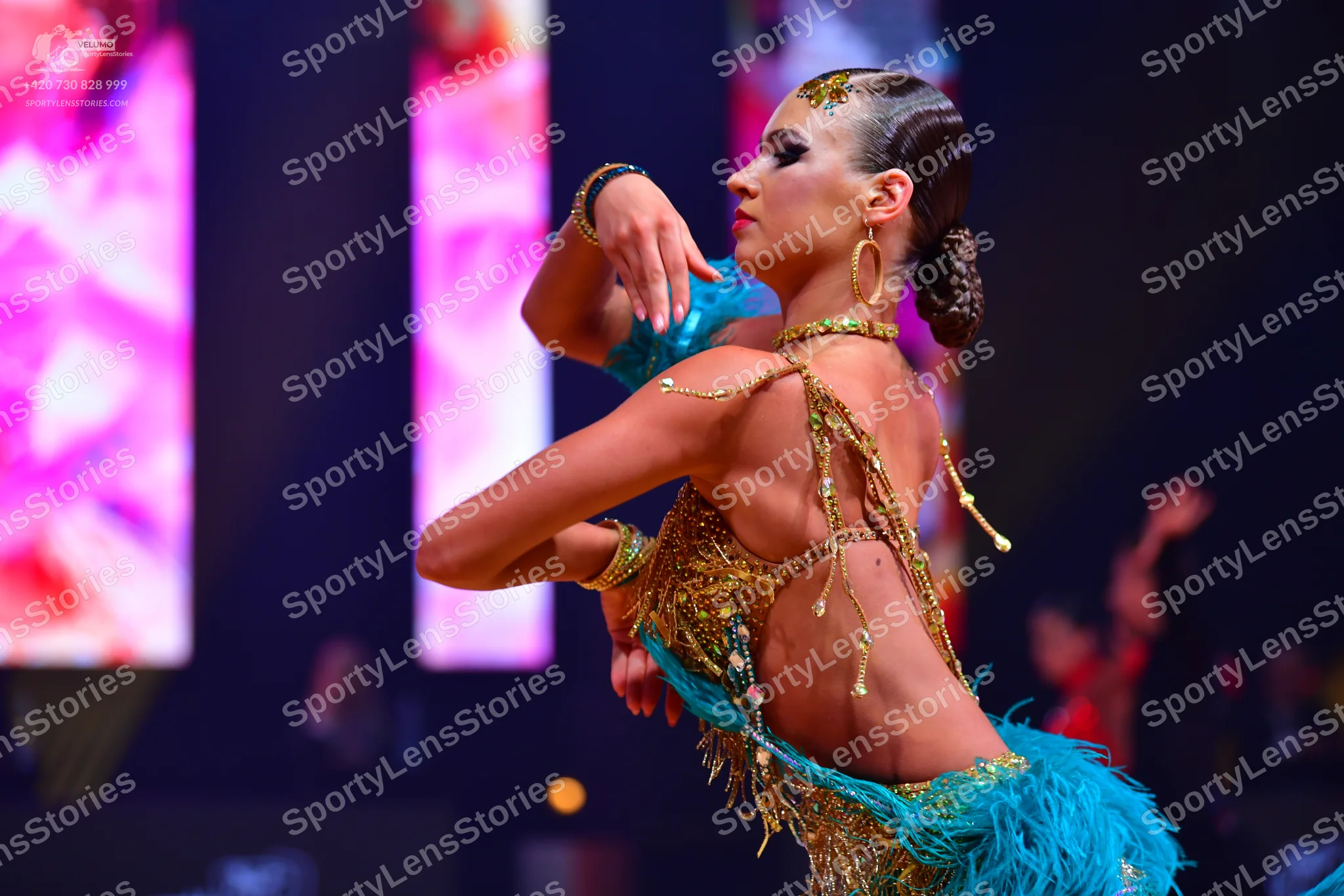Dancesport competitor: Female dancer: Gold sequined Latin costume with teal feather and fringe accents,