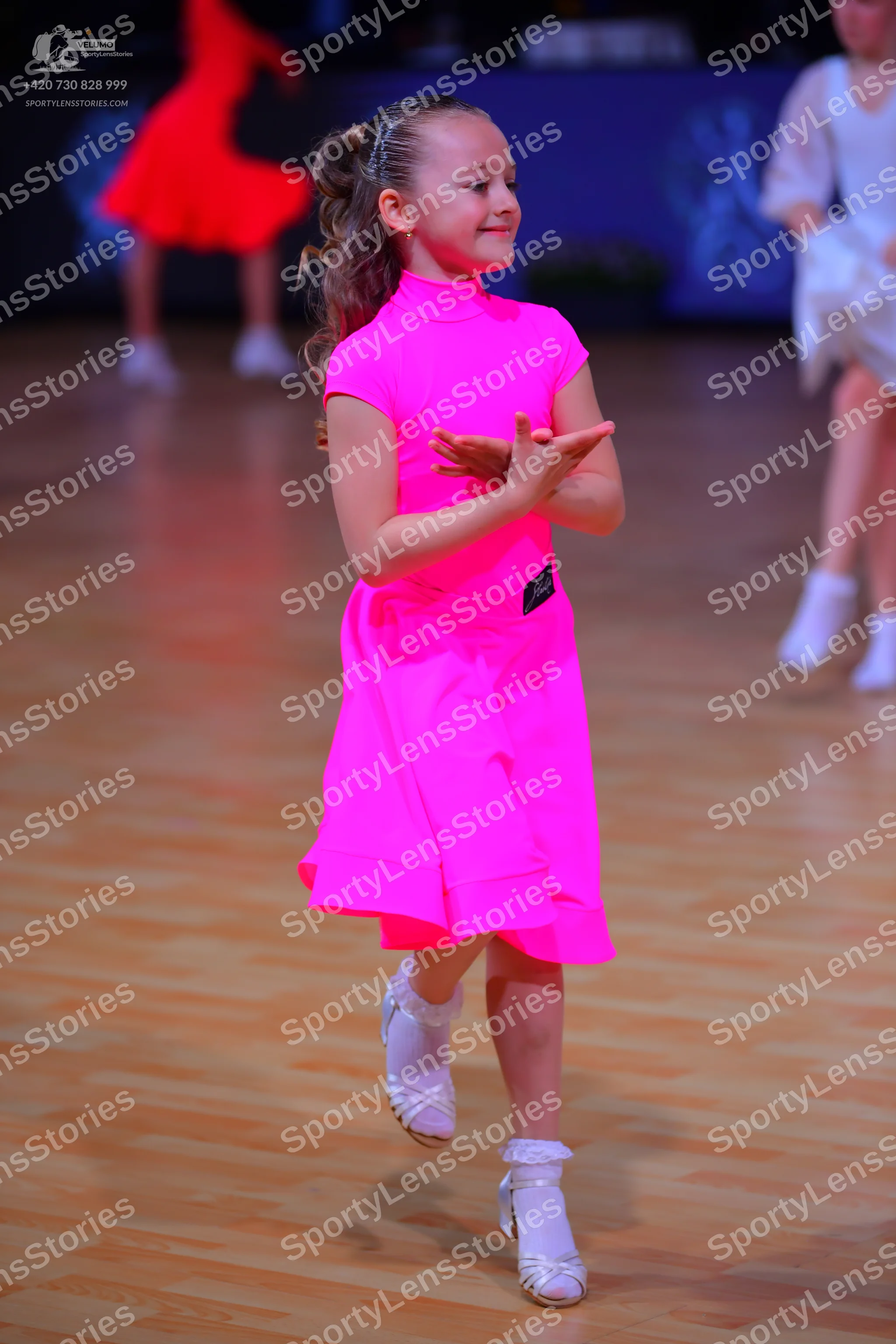 Dancesport competitor: Female dancer in foreground: bright pink short-sleeved dress with a high neck an