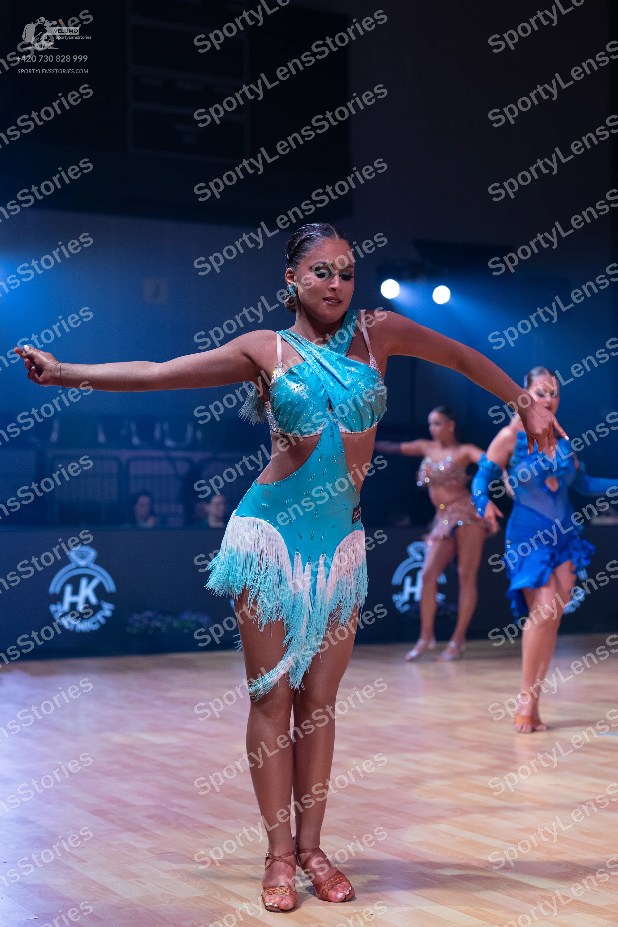 Dancesport competitor: Female dancer in foreground: teal latin dance dress with fringe and cutouts