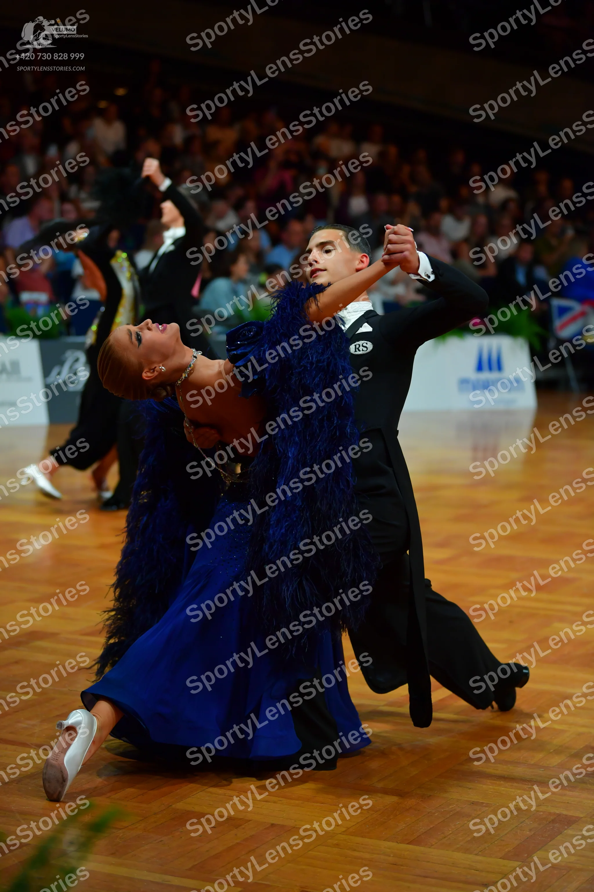Dancesport competitor: Foreground female dancer: deep blue ballgown with large dark blue feathery/fring