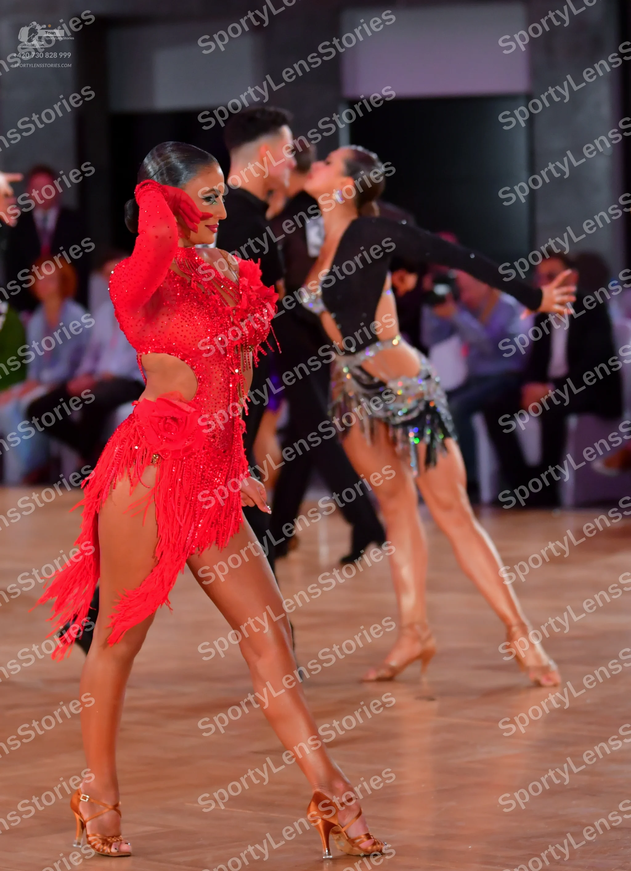 Dancesport competitor: Female dancer in foreground: red Latin dance dress with long sleeves, cutouts, a