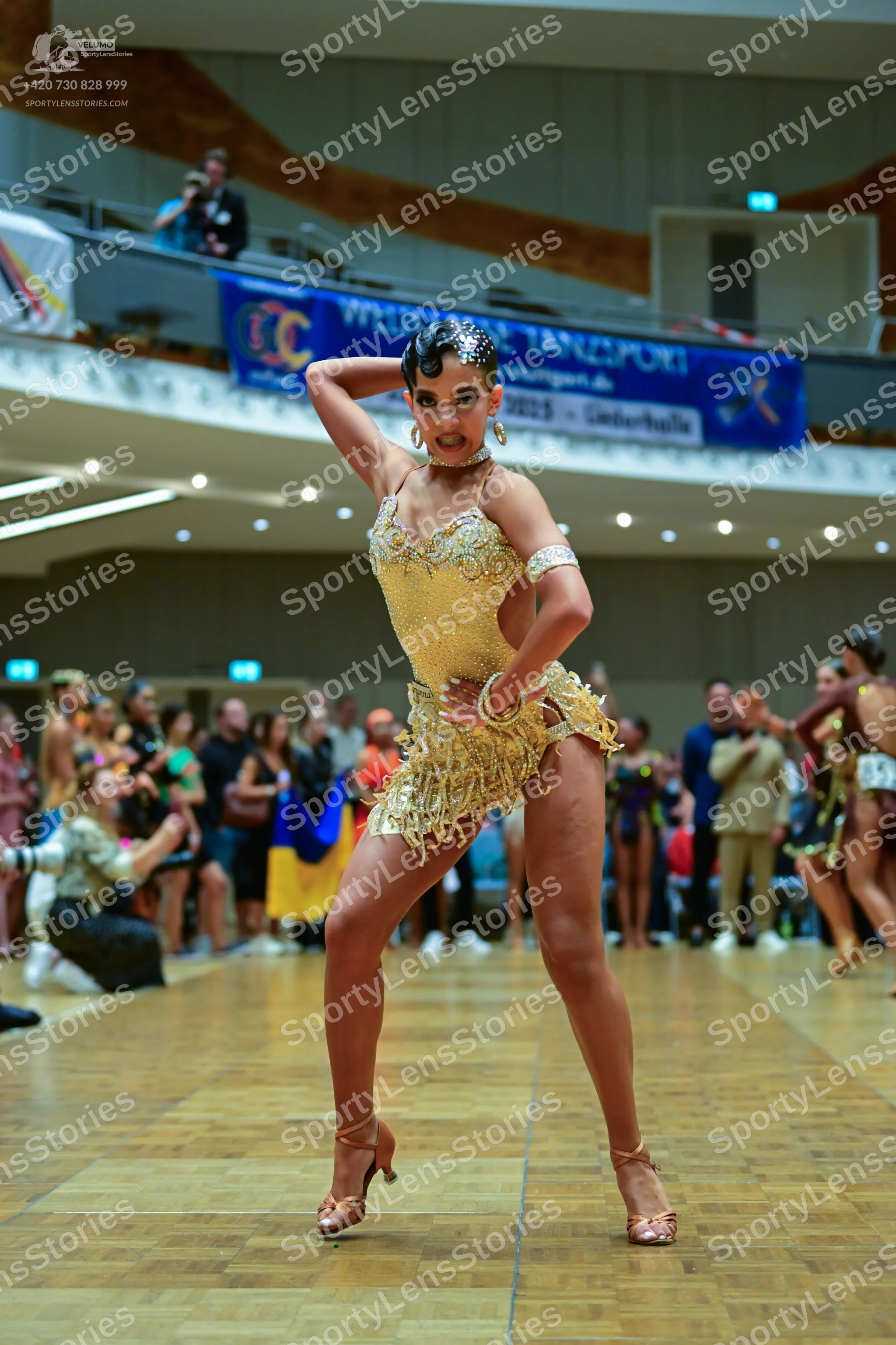 Dancesport competitor: Female dancer in foreground: champagne-colored Latin dance dress heavily embelli