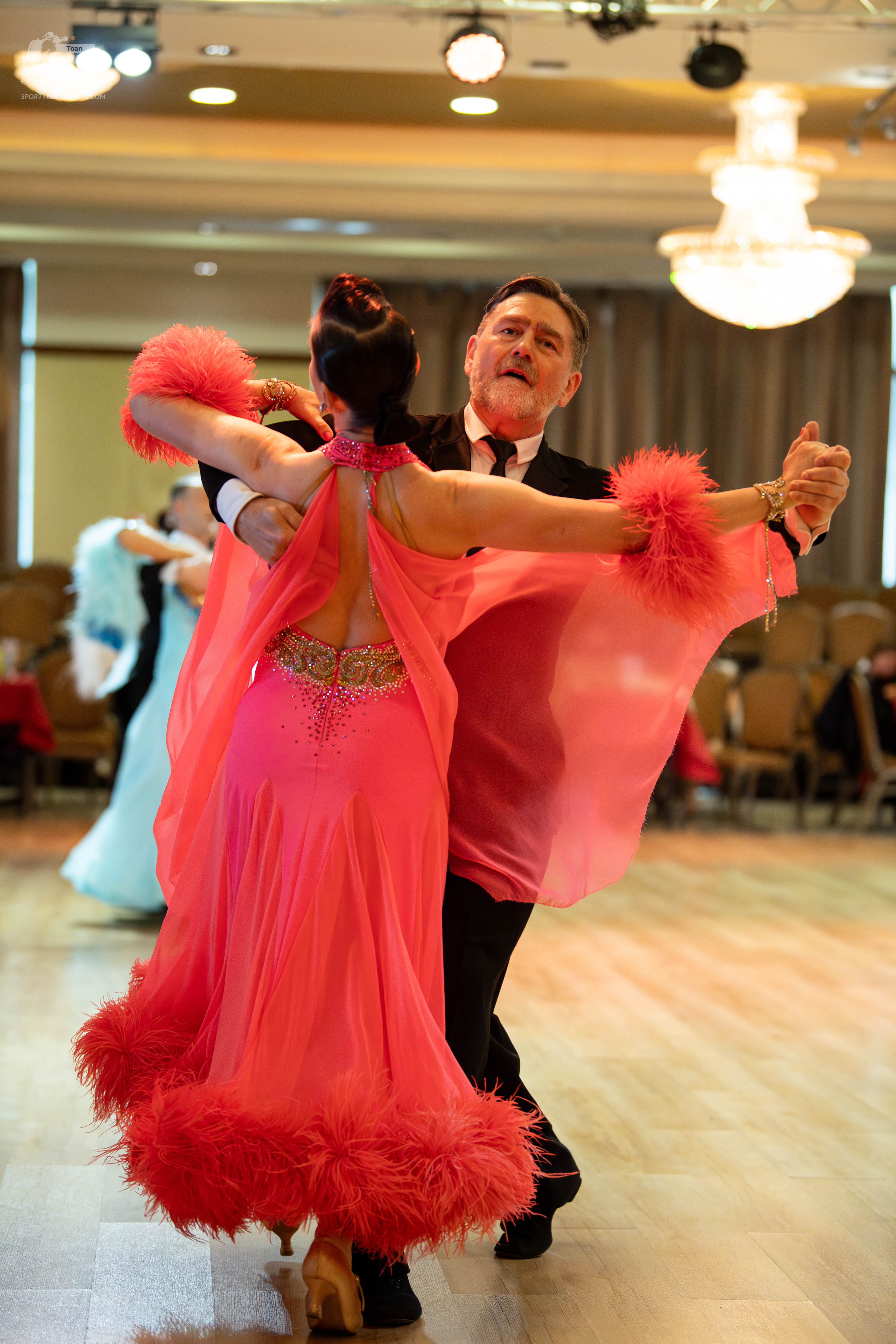 Dancesport competitor: Female dancer in foreground: bright pink/coral dress with feather trim on the sl
