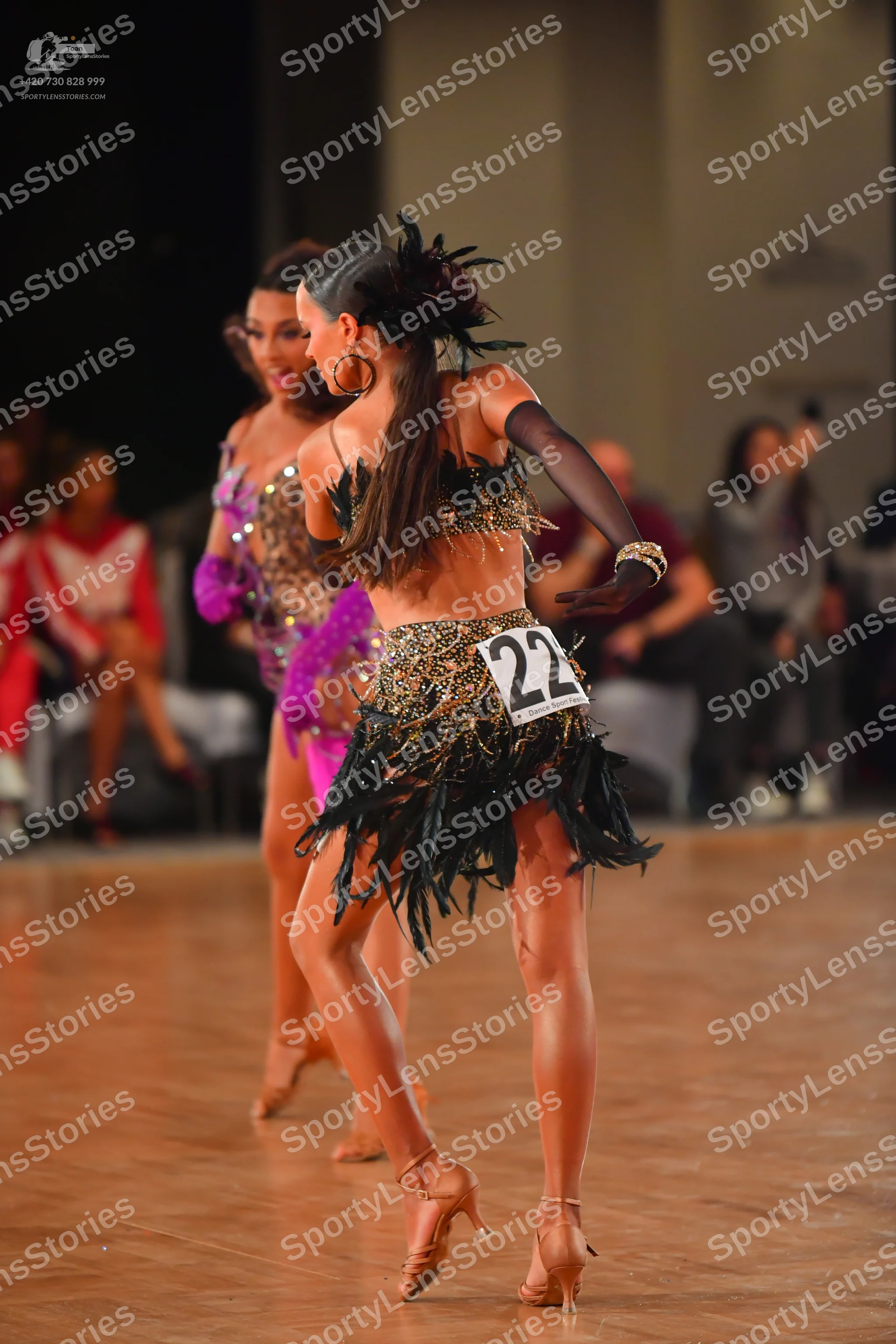 Dancesport competitor #22: Female dancer in foreground: black and gold sequin Latin dress with a feathered/