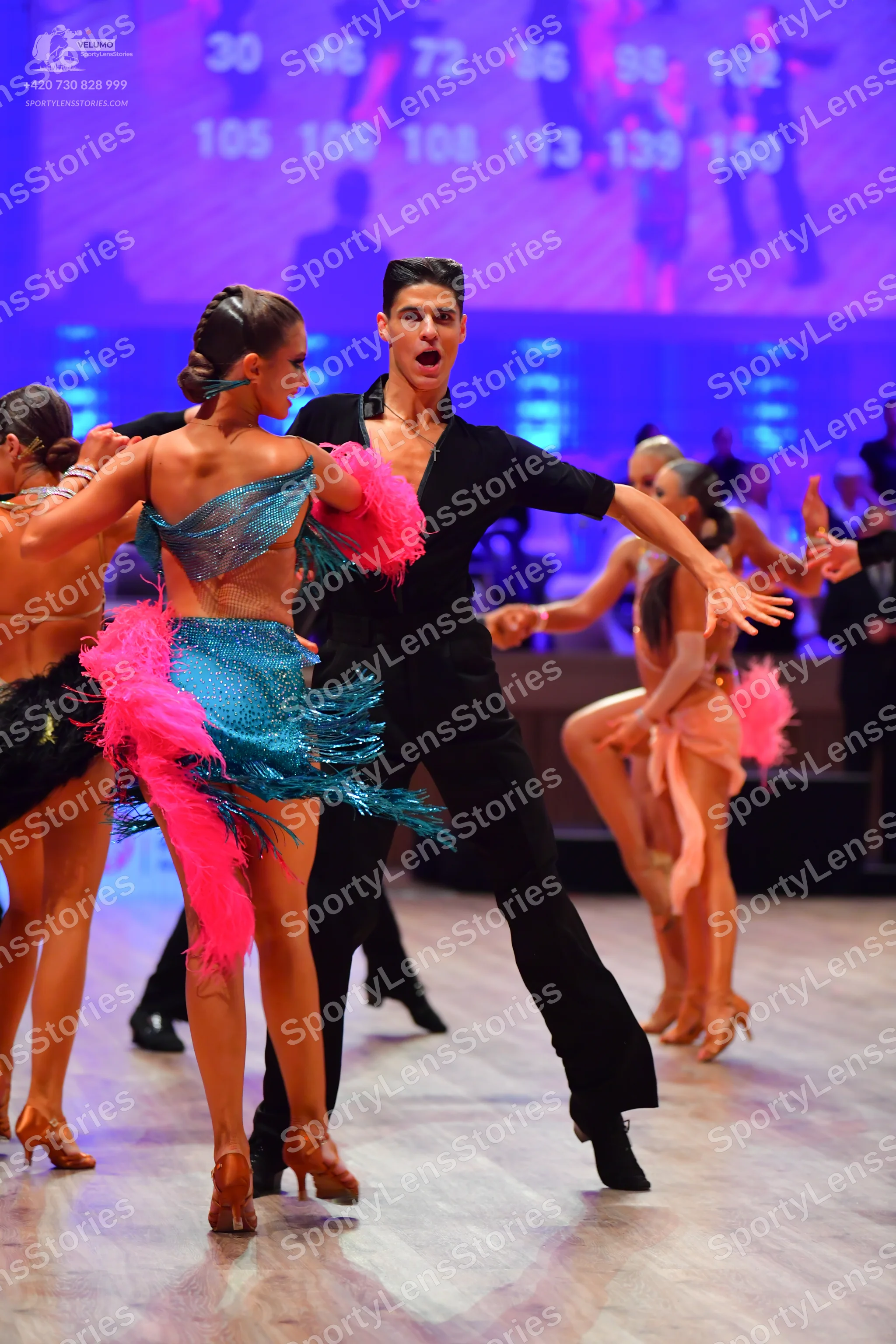 Dancesport competitor: Female dancer in foreground: teal sequined latin dress with bright pink feather-