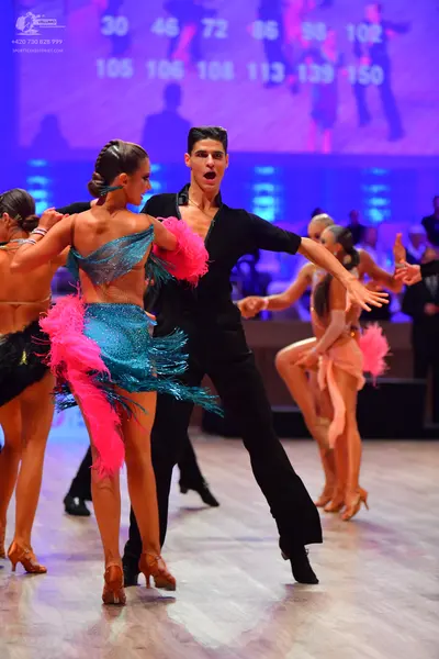 Dancesport competitor: Female dancer in foreground: teal sequined latin dress with bright pink feather-