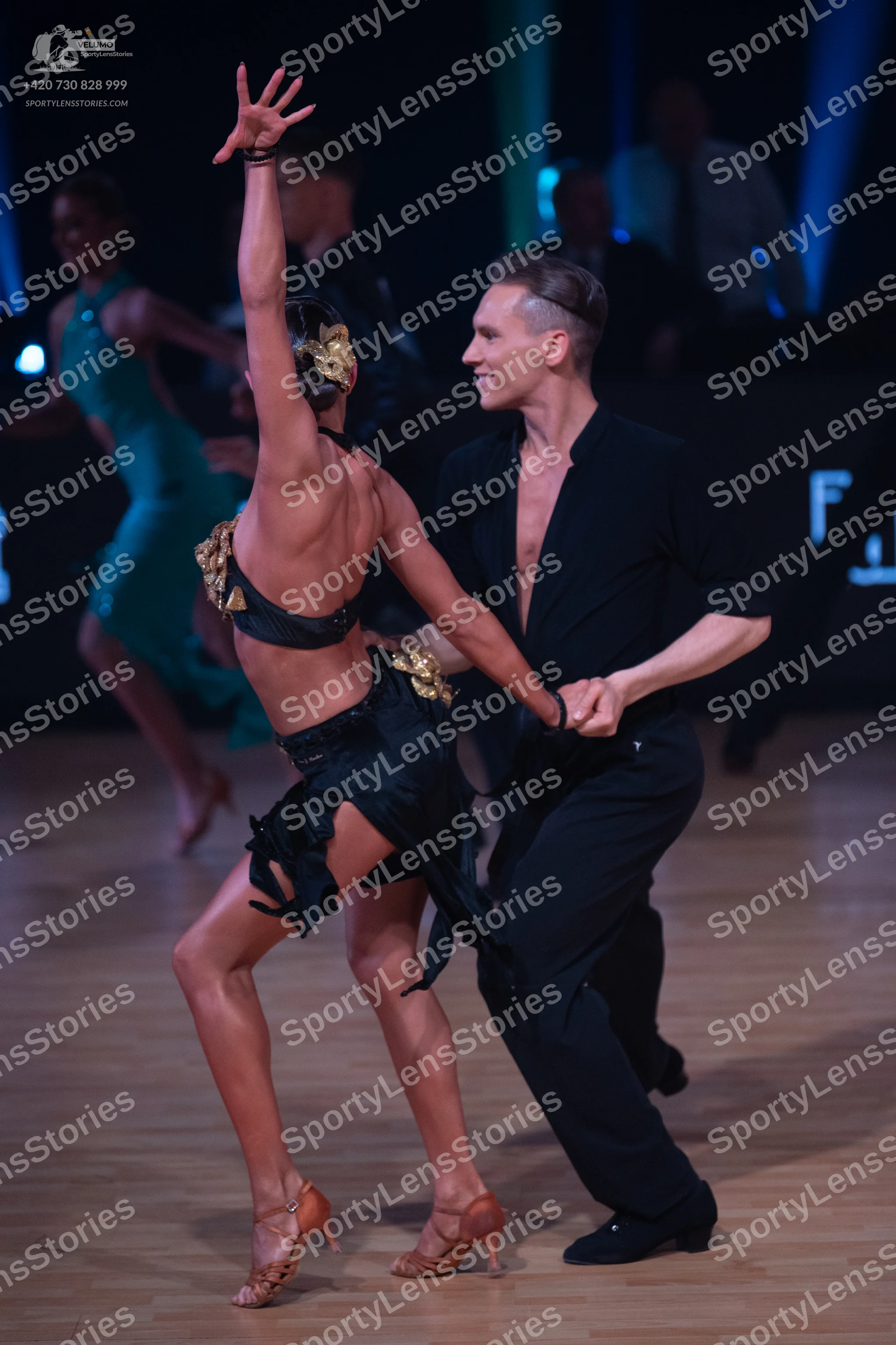 Dancesport competitor: Female dancer in foreground: black backless Latin dance costume with gold embell