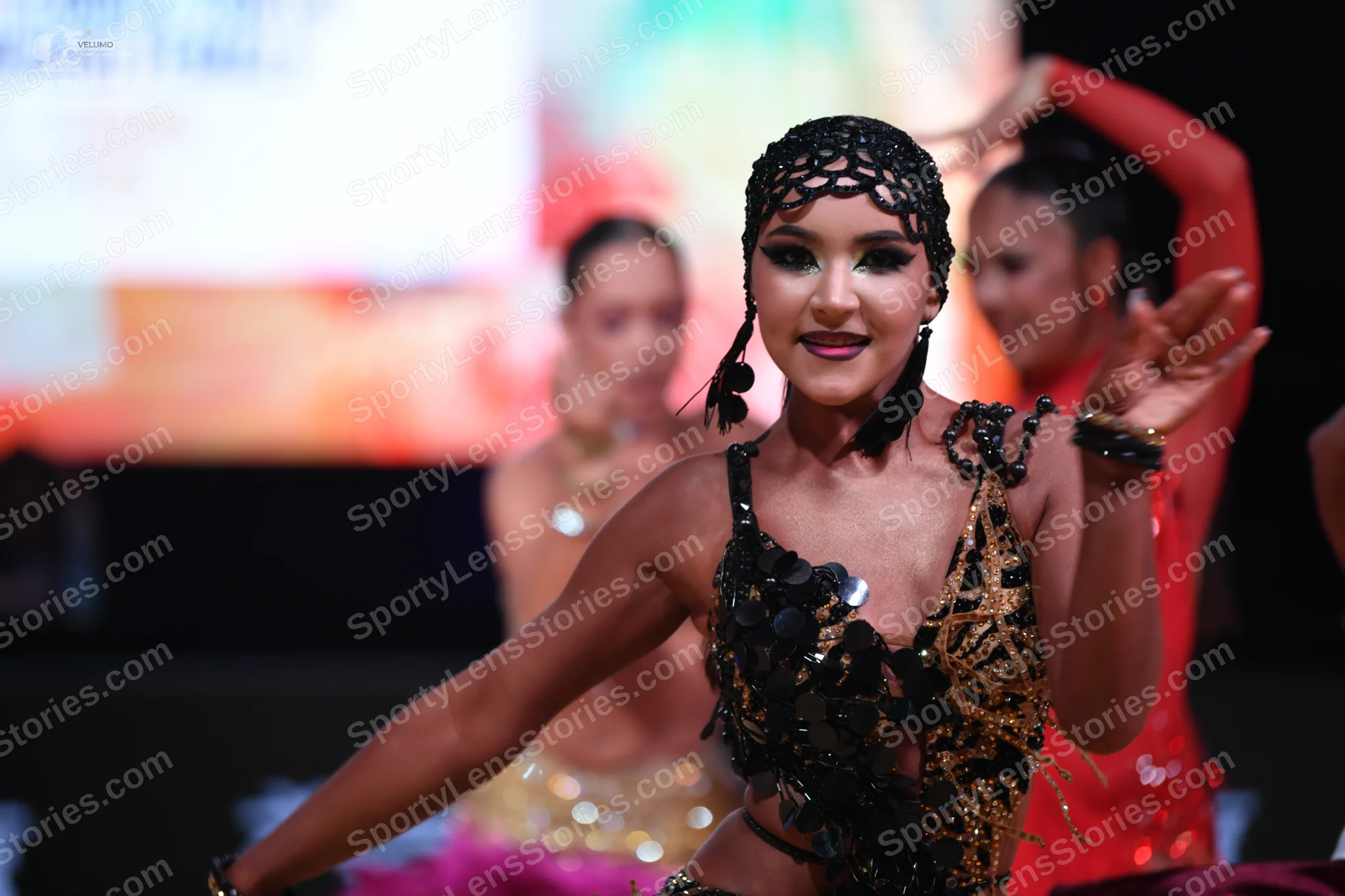 Dancesport competitor: Foreground dancer: black Latin costume with gold/bronze sequins, large dark circ