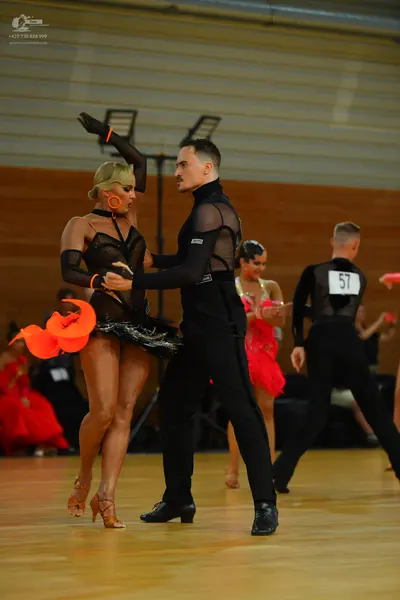Dancesport competitor: Foreground female dancer: black latin dress with sheer mesh sleeves and large or