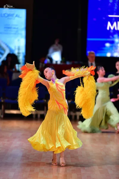 Dancesport competitor: Female dancer in foreground: bright yellow Latin dress featuring large, fluffy y