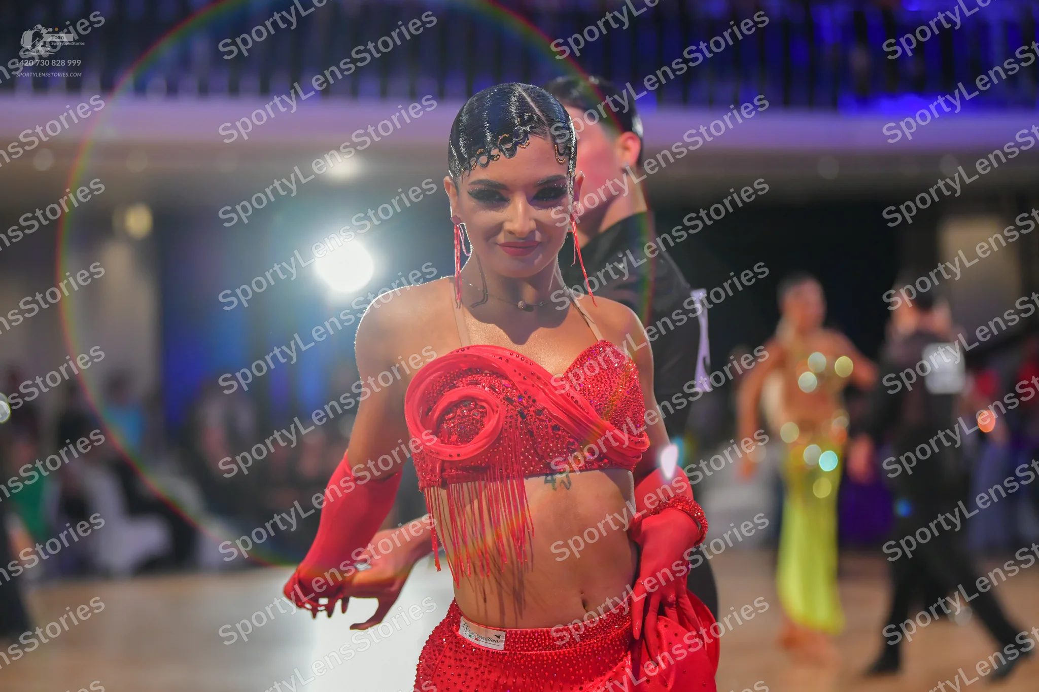 Dancesport competitor: Female dancer in foreground: red latin dress with sequins and fringe, and long r