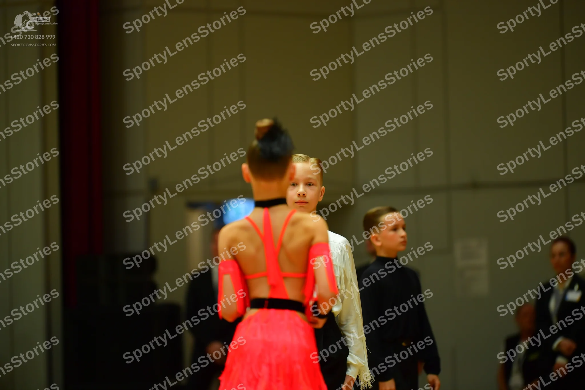 Dancesport competitor: Foreground dancer: bright orange Latin dress with a black waistband and sheer me