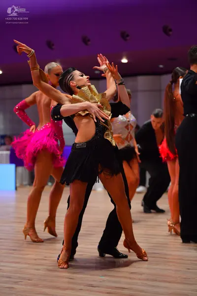 Dancesport competitor: Female dancer in foreground: black latin dress with a large gold sequined collar