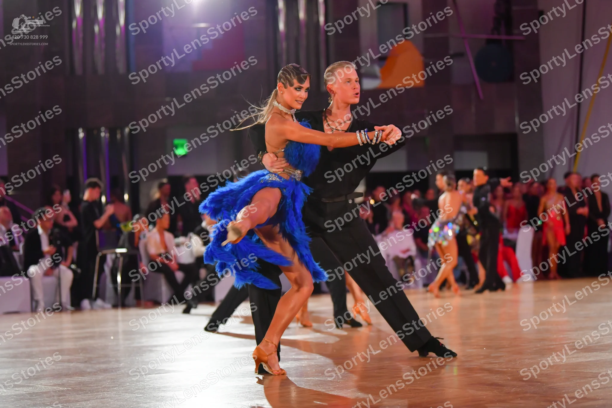 Dancesport competitor: Foreground female dancer: blue Latin dress with fringe and sequins, paired with 