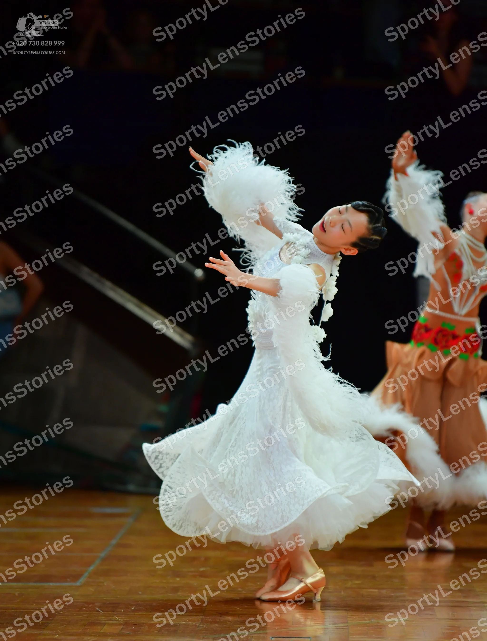 Dancesport competitor: Female dancer in foreground: white ballroom dress featuring lace, a voluminous s