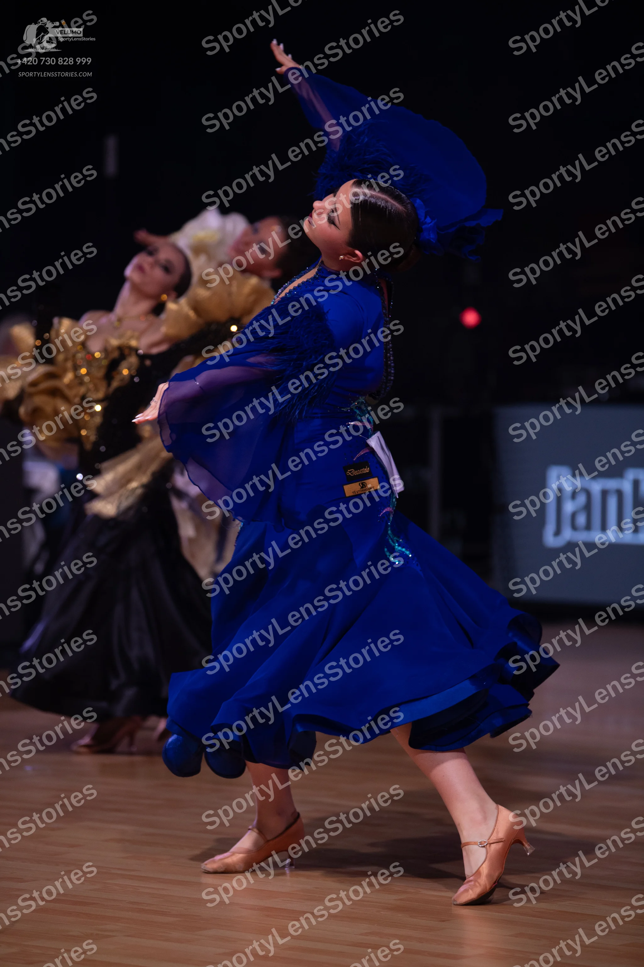 Dancesport competitor: Female dancer in foreground: royal blue ballroom gown with large, flowing, sheer