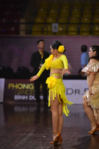 Dancesport competitor: Female dancer: yellow latin dress with long sleeves and fringe, yellow flower in