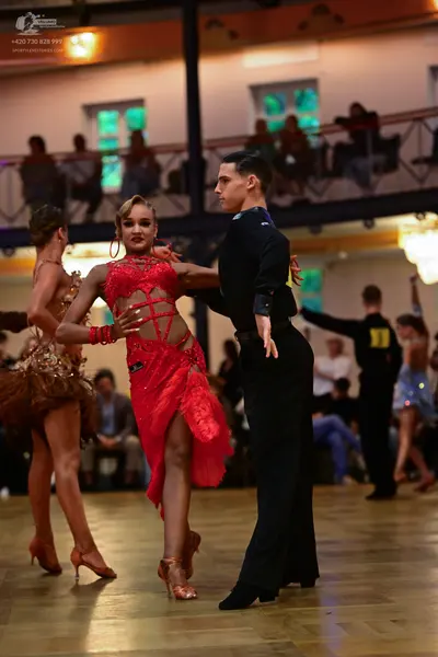 Dancesport competitor: Female dancer in foreground: red lace Latin dress with cutouts and a slit, paire