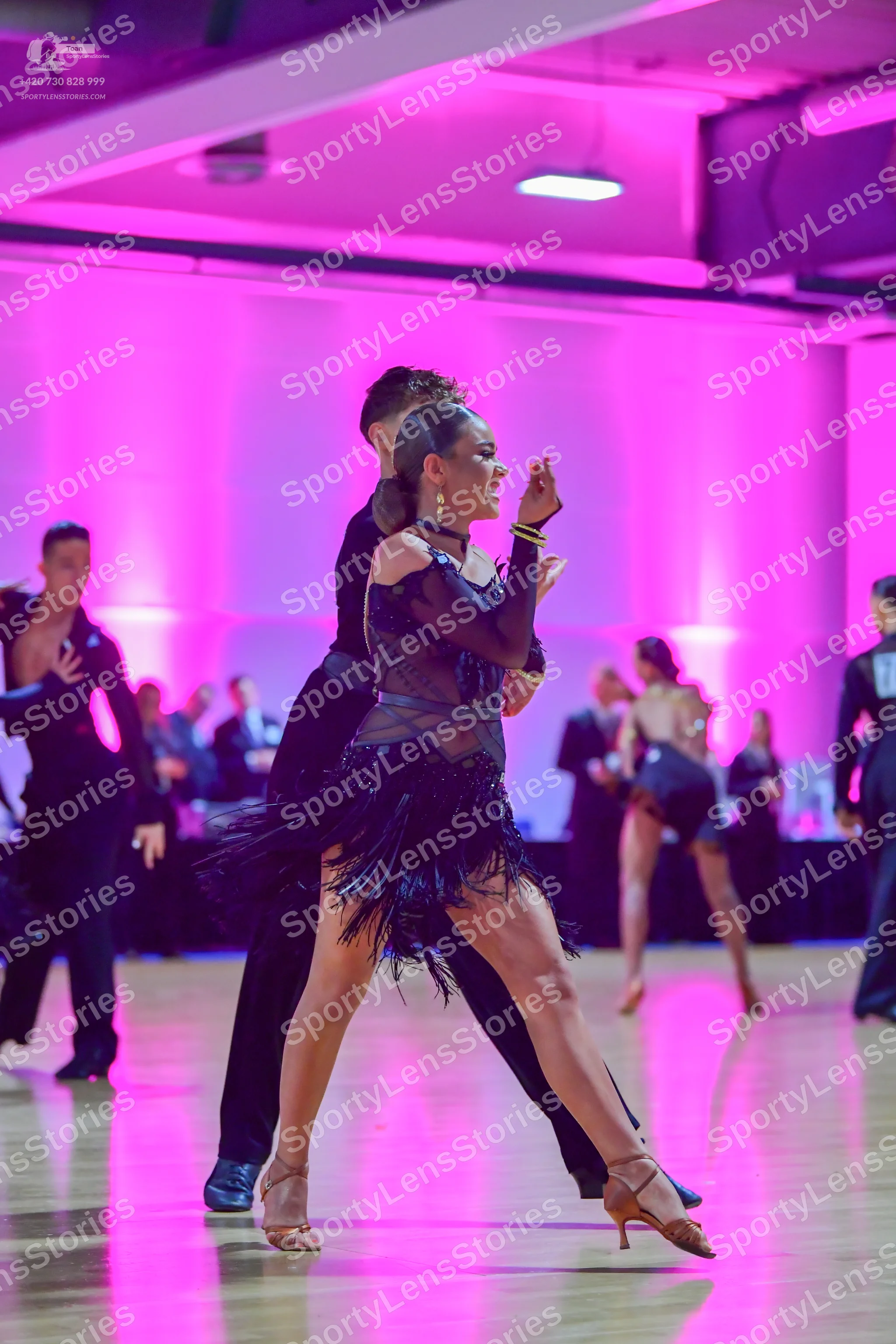 Dancesport competitor: Foreground female dancer: black latin dress with a sheer mesh bodice and a black