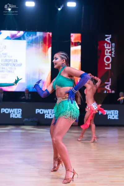 Dancesport competitor: Female dancer in foreground: teal latin dance dress with fringe, a gold waistban