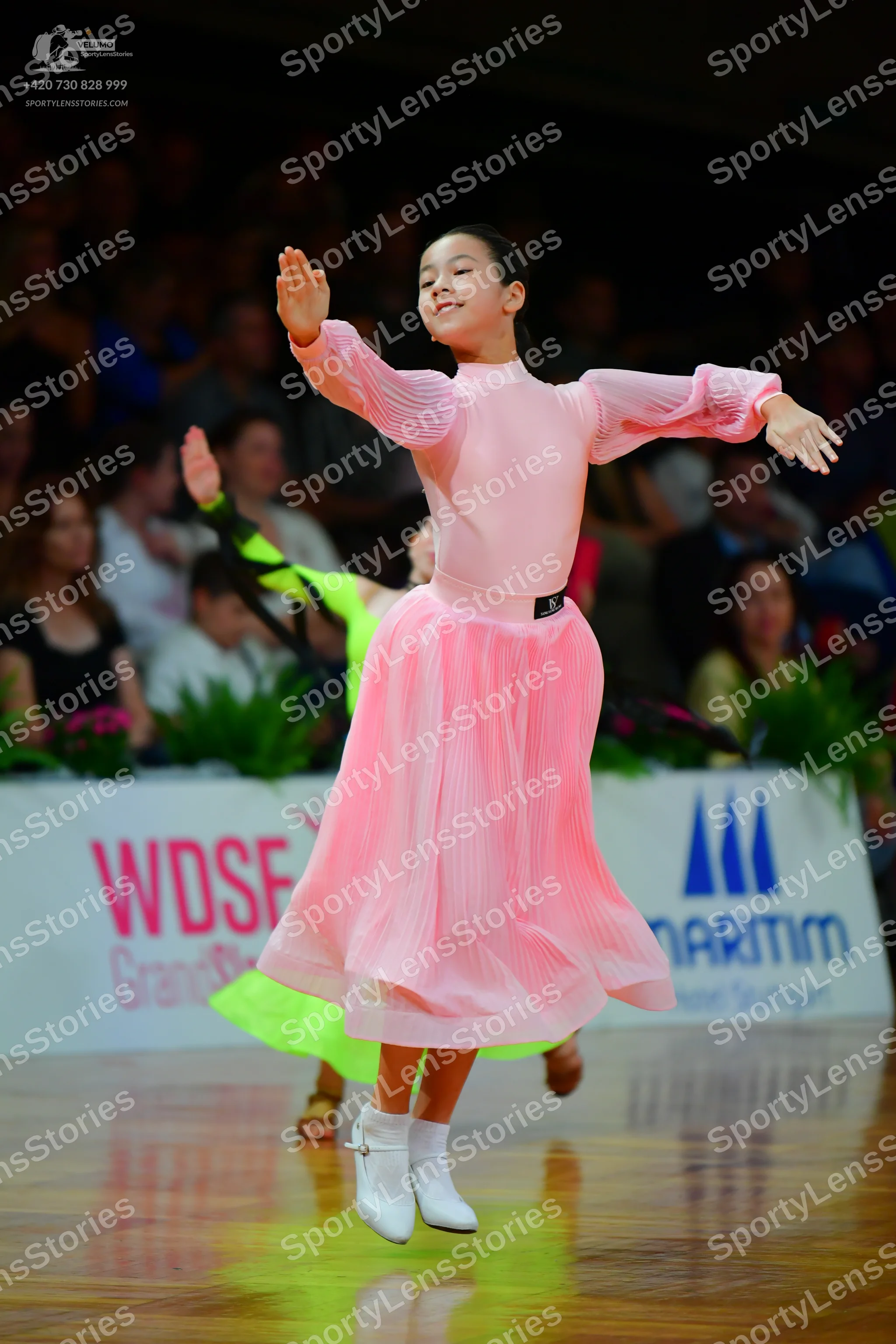 Dancesport competitor: Female dancer in foreground: light pink long-sleeved dress with pleated sleeves