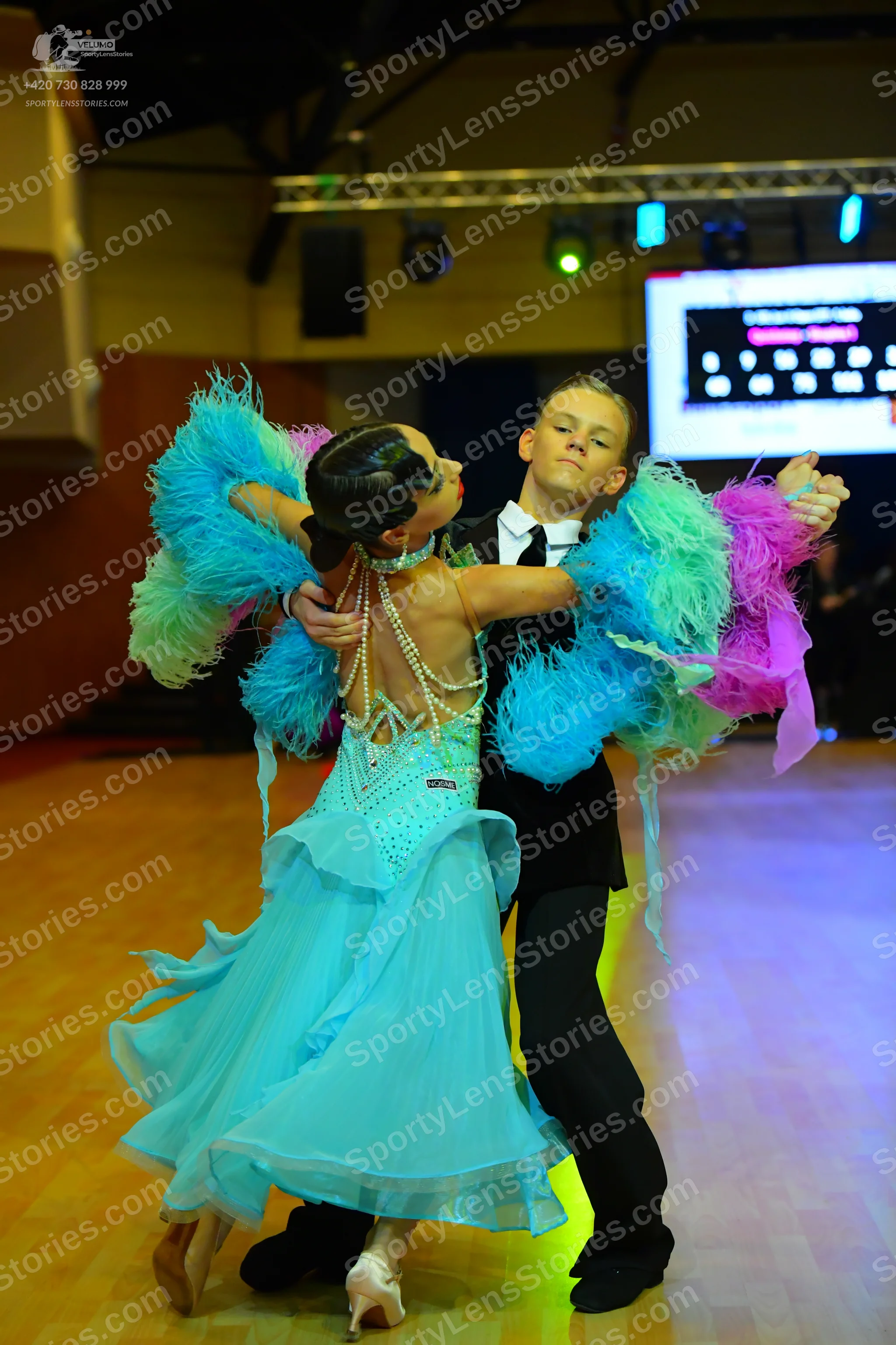 Dancesport competitor: Female dancer: light blue/aqua dance dress with large, colorful feathers (blue,